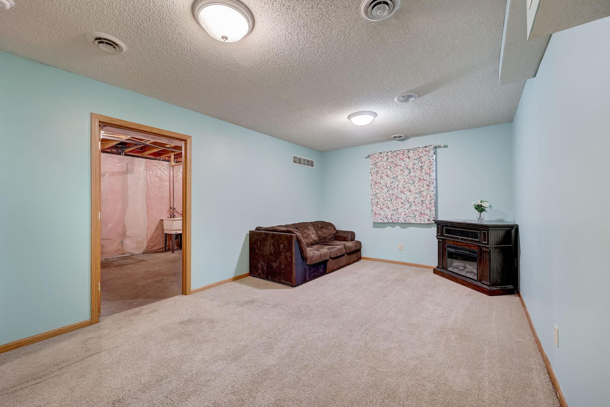 Basement living room with a brown sofa and corner fireplace beside an unfinished adjoining area.
