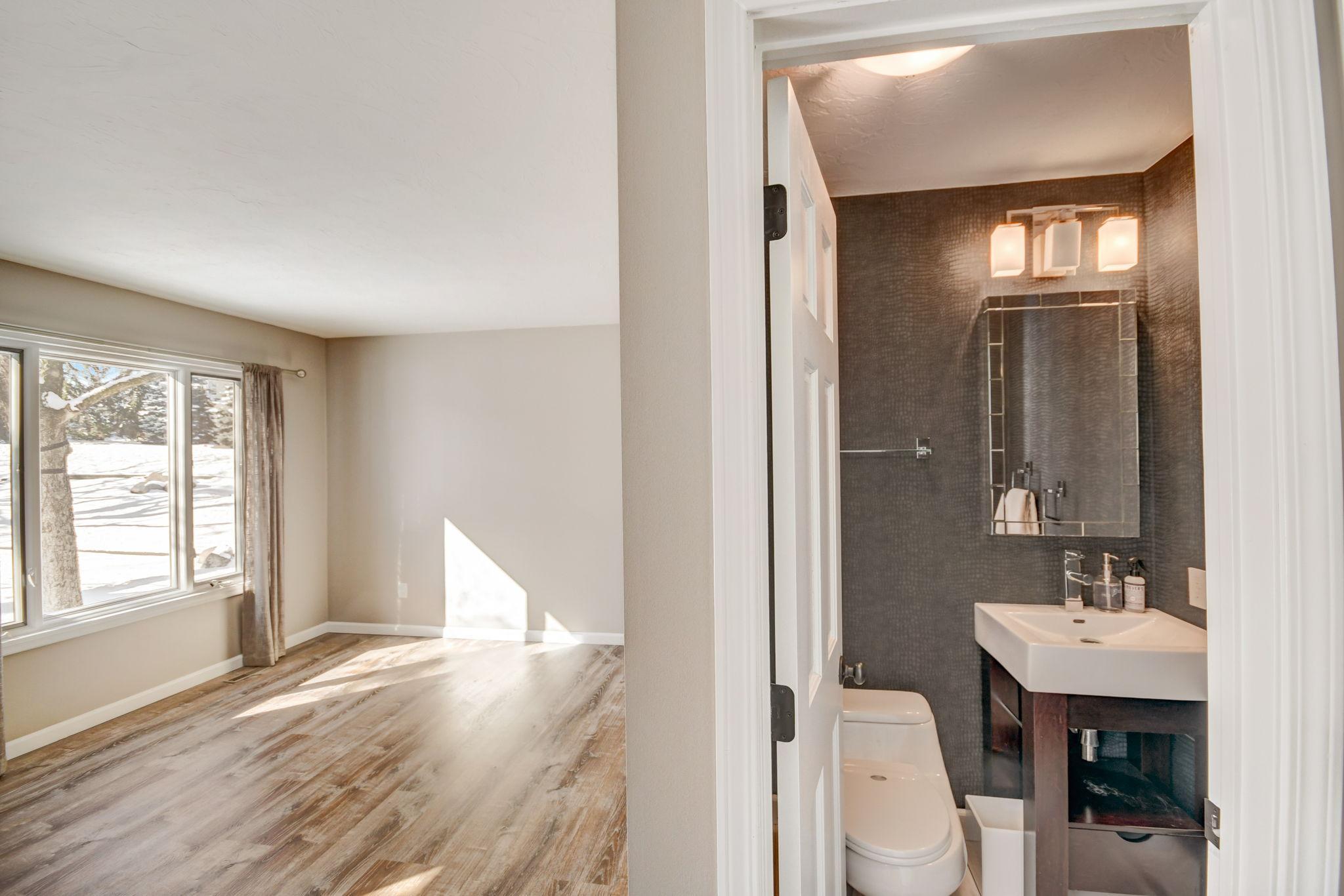 Empty bedroom with wood-look flooring and snowy views beside a bathroom with a dark tiled wall and white vanity.