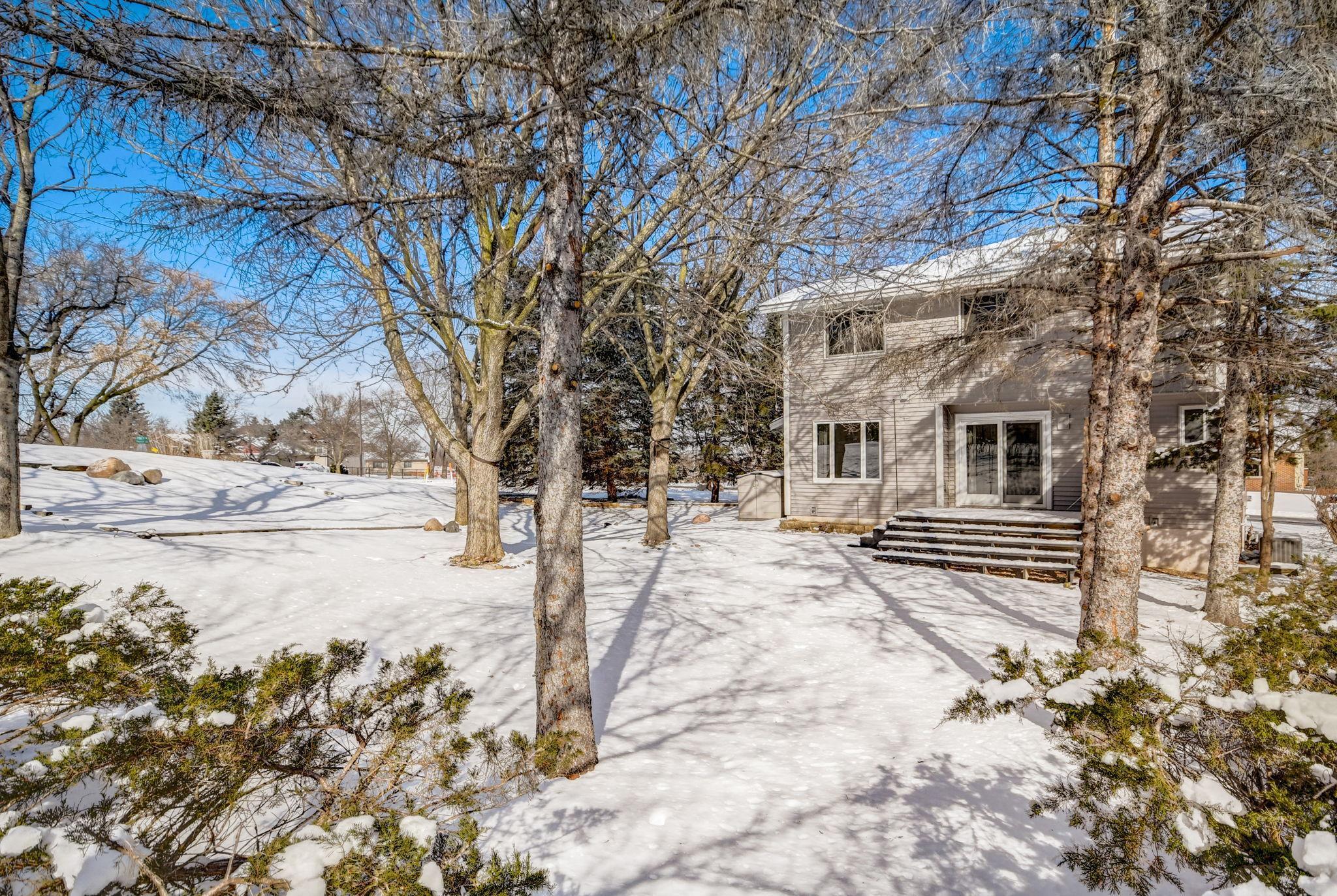 Rear of a gray two-story house with patio doors and steps in a snowy, tree-filled yard.