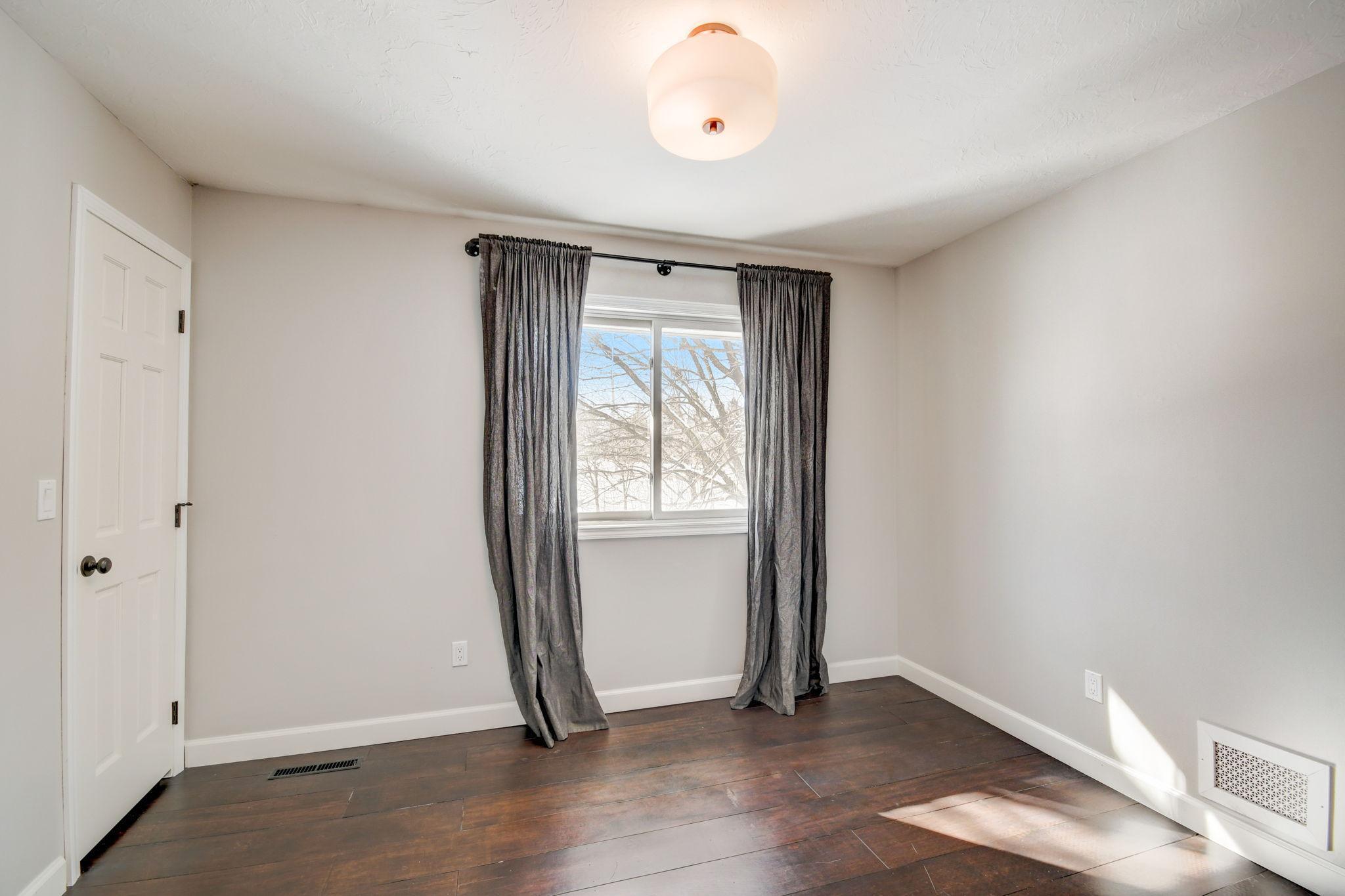 Vacant bedroom with dark wood floors, gray curtains, and sunlight through the window.