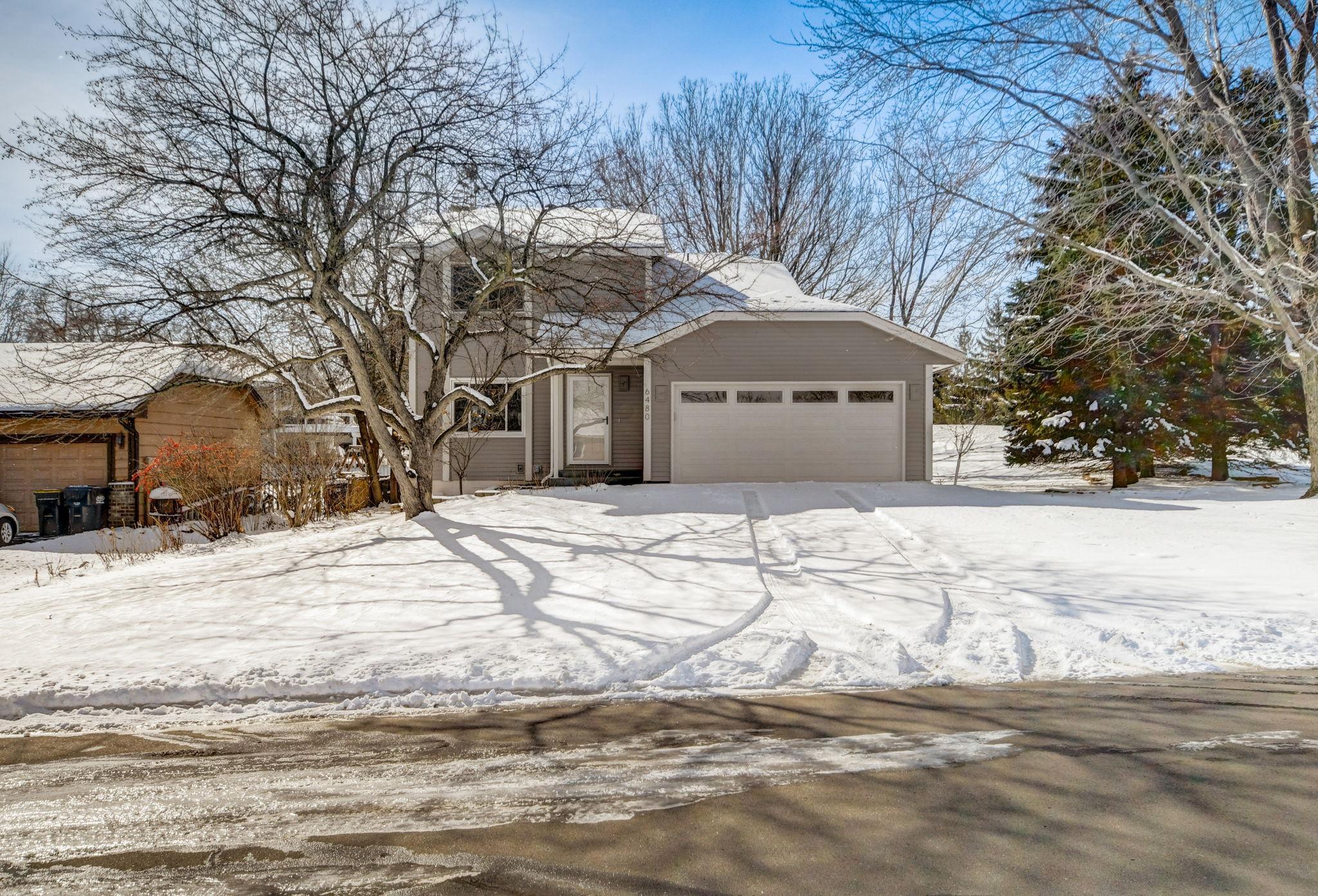 Gray split-level home with an attached garage, snow-covered yard, and leafless trees.