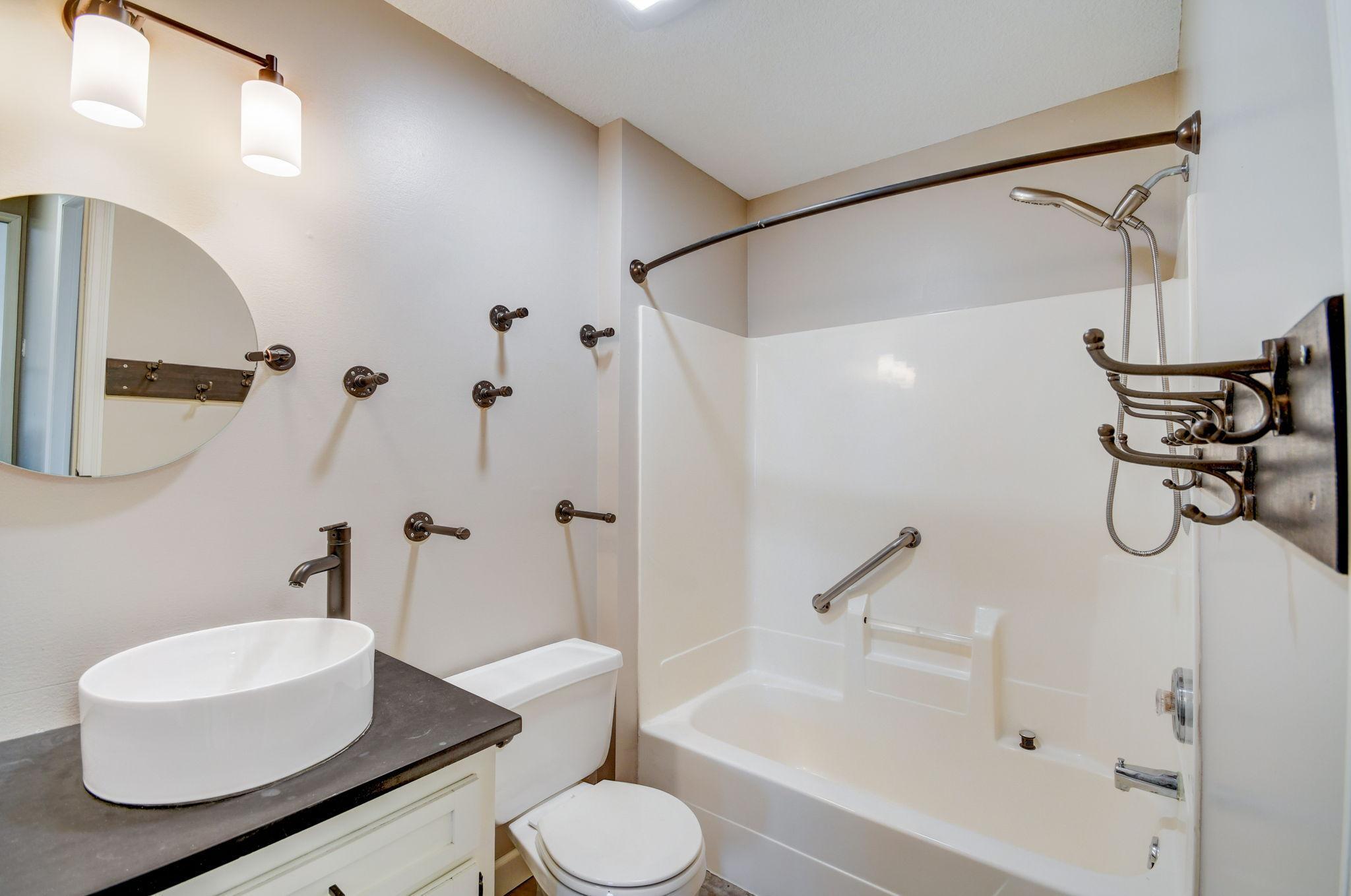 Bathroom with a vessel sink, round mirror, toilet, and bathtub with shower and grab bars.