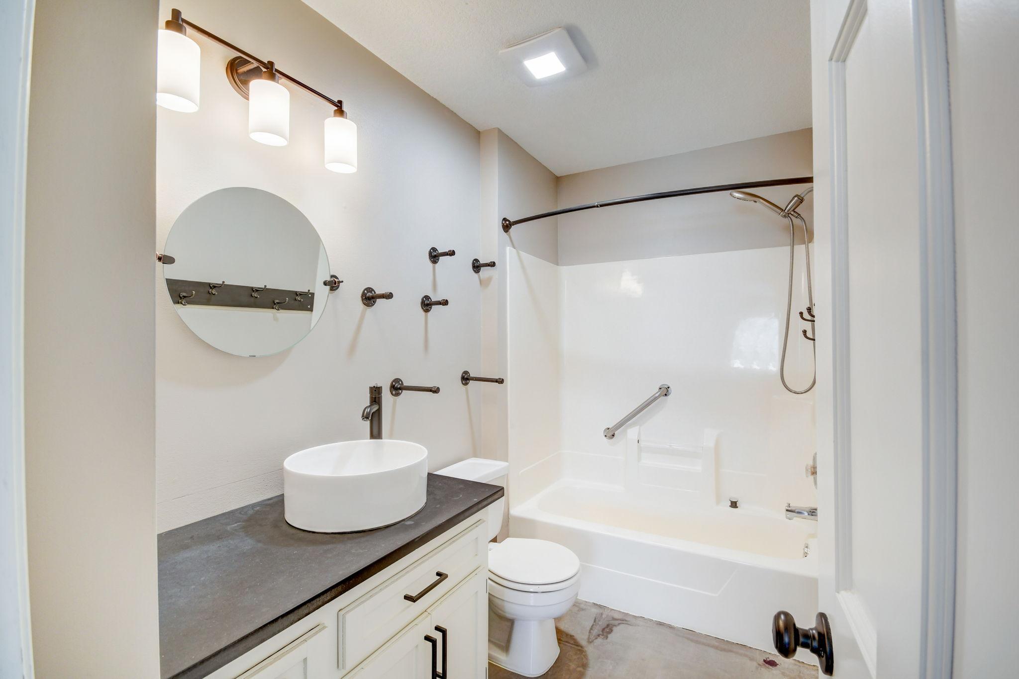 Bathroom with vessel sink and round mirror beside a bathtub with grab bars and handheld shower.