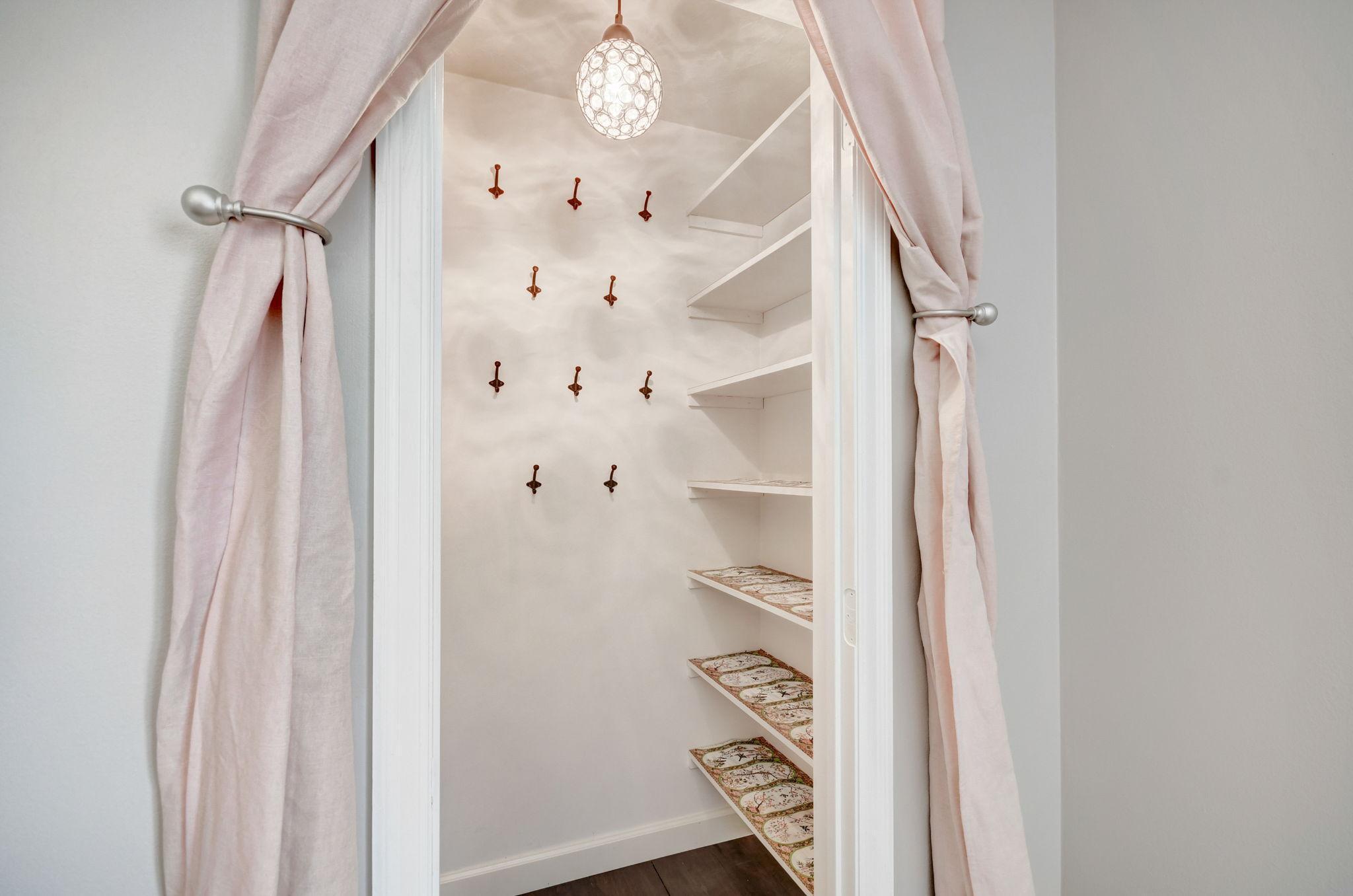 Walk-in closet with wall hooks, patterned shelf liners, and pale pink curtains framing the doorway.