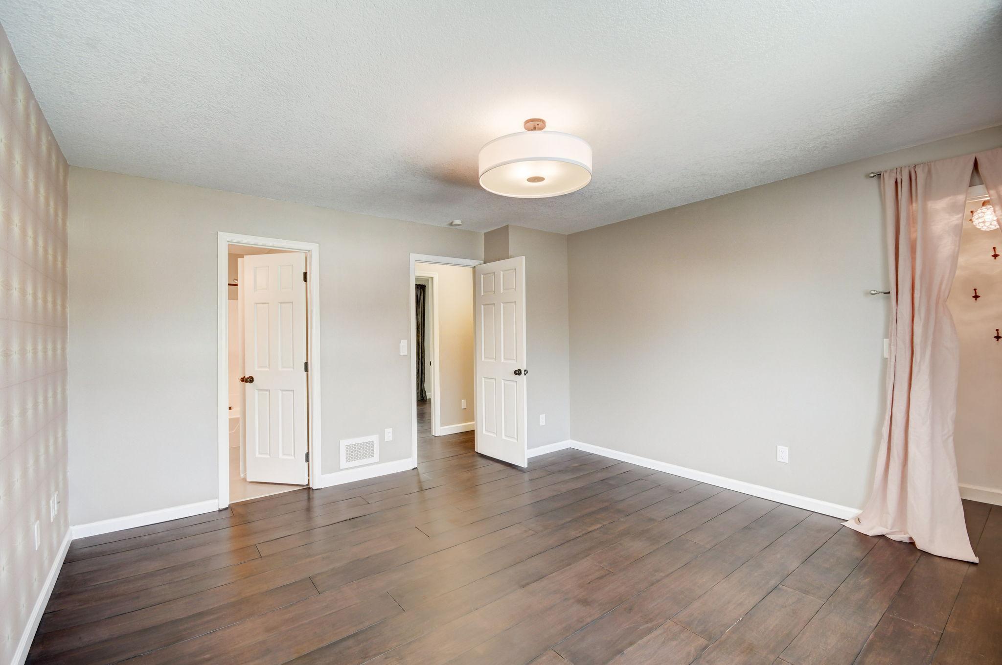 Empty bedroom with plank flooring, two open doors, and pale pink curtains.