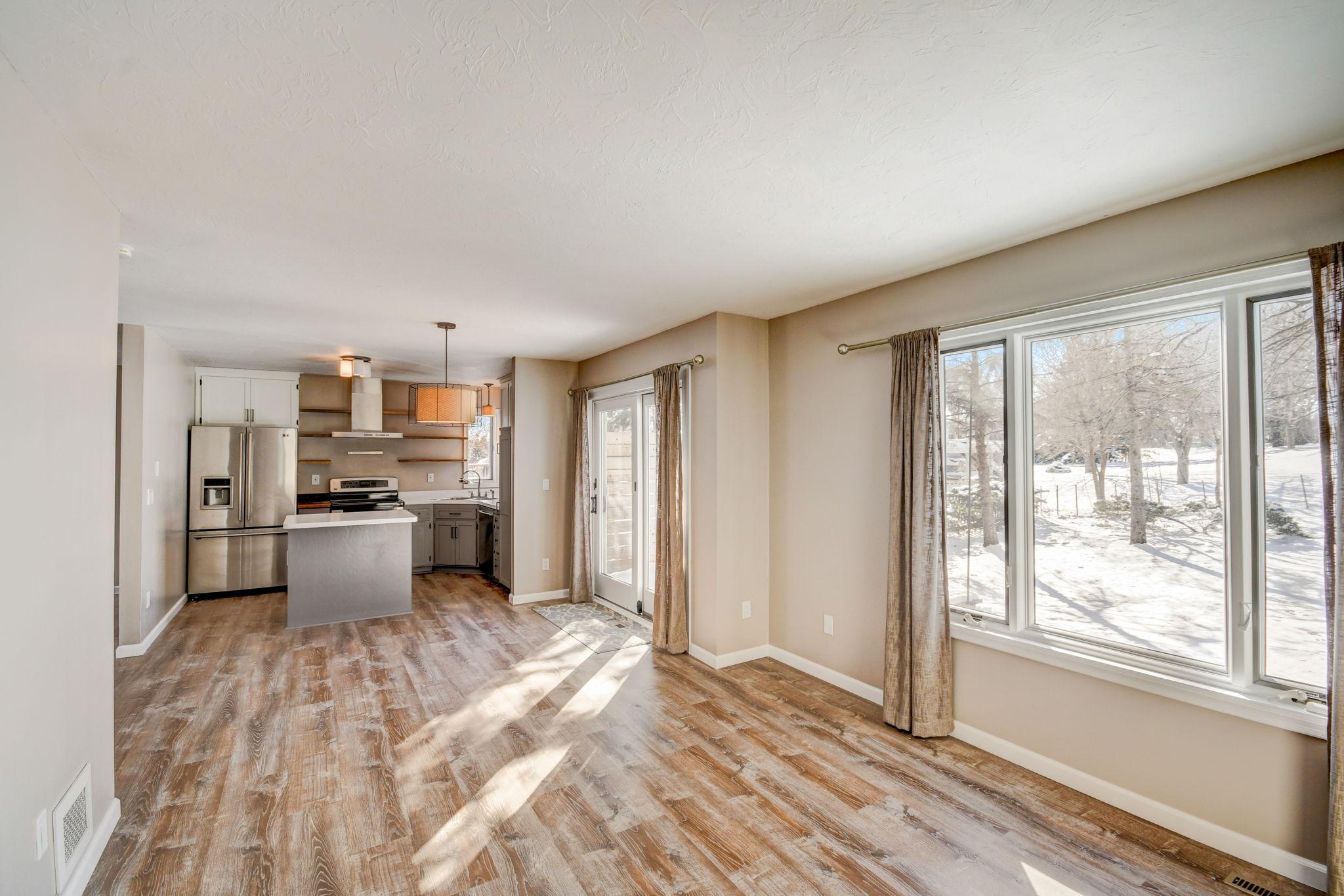 Open living area and kitchen with wood-look floors, stainless appliances, and large windows overlooking a snowy yard.