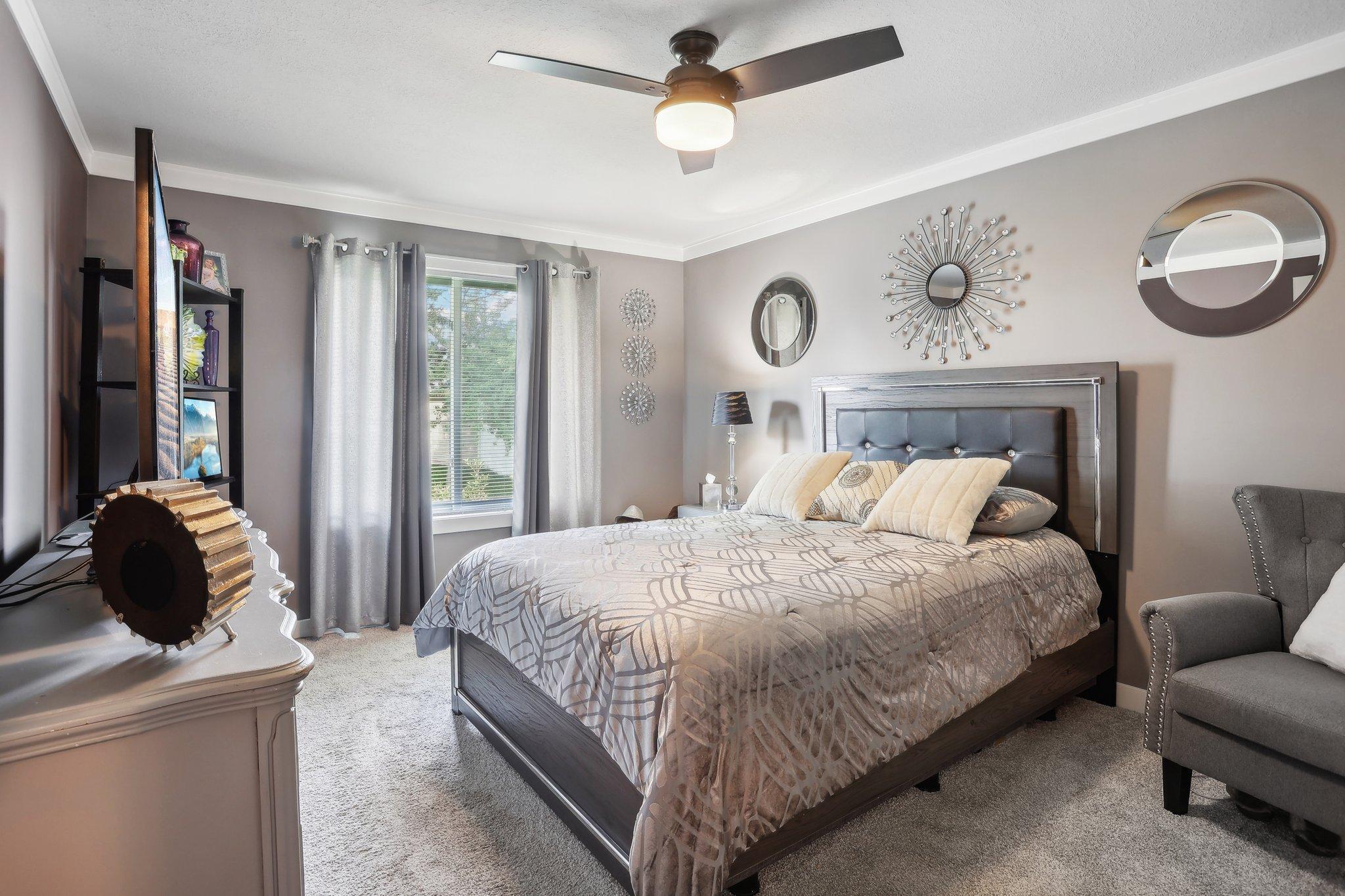 Furnished bedroom with a large gray bed, ceiling fan, decorative mirrors, and an armchair.