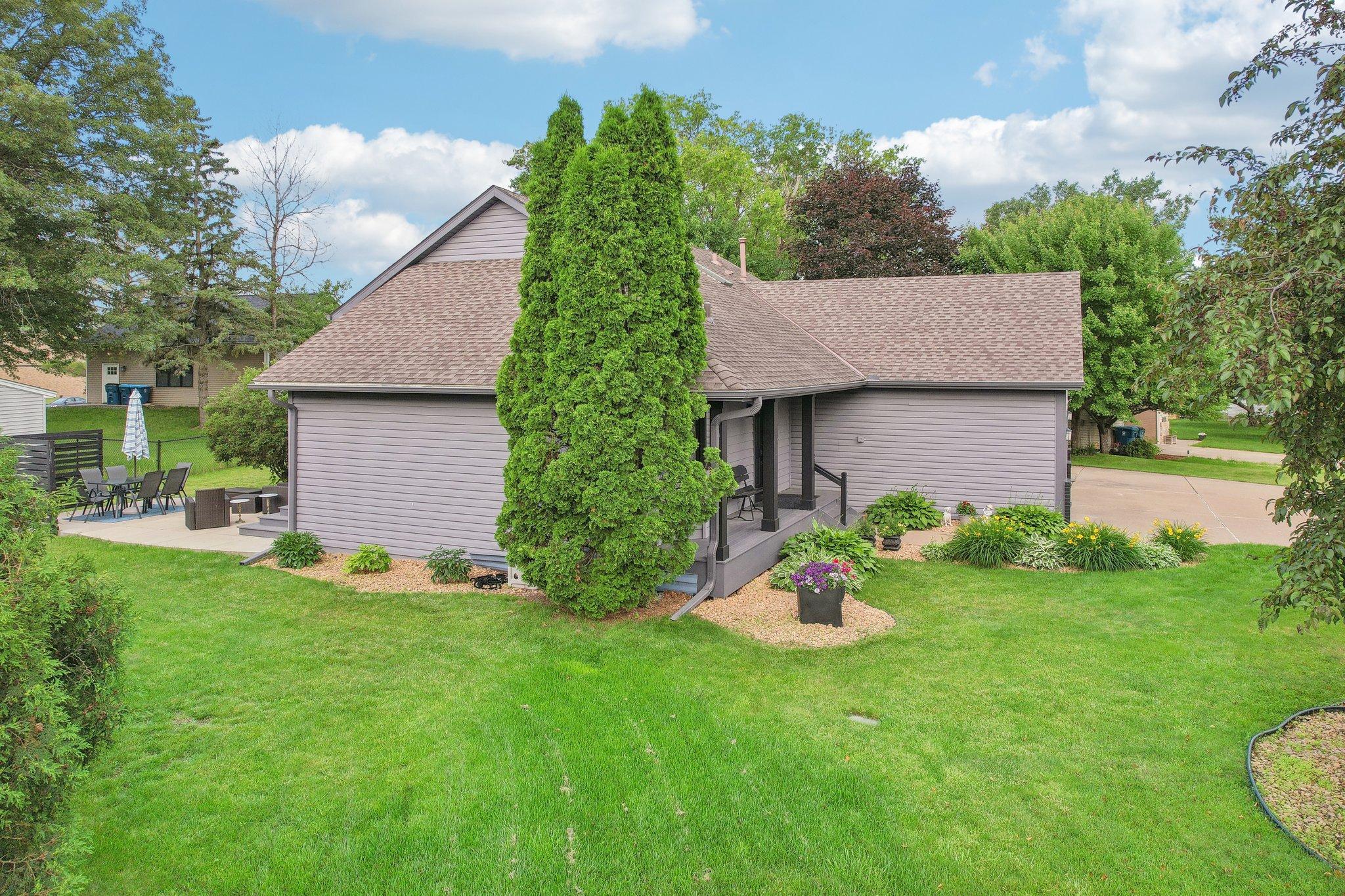 Gray-sided house with a covered porch, patio seating, and a tall evergreen in the lawn.