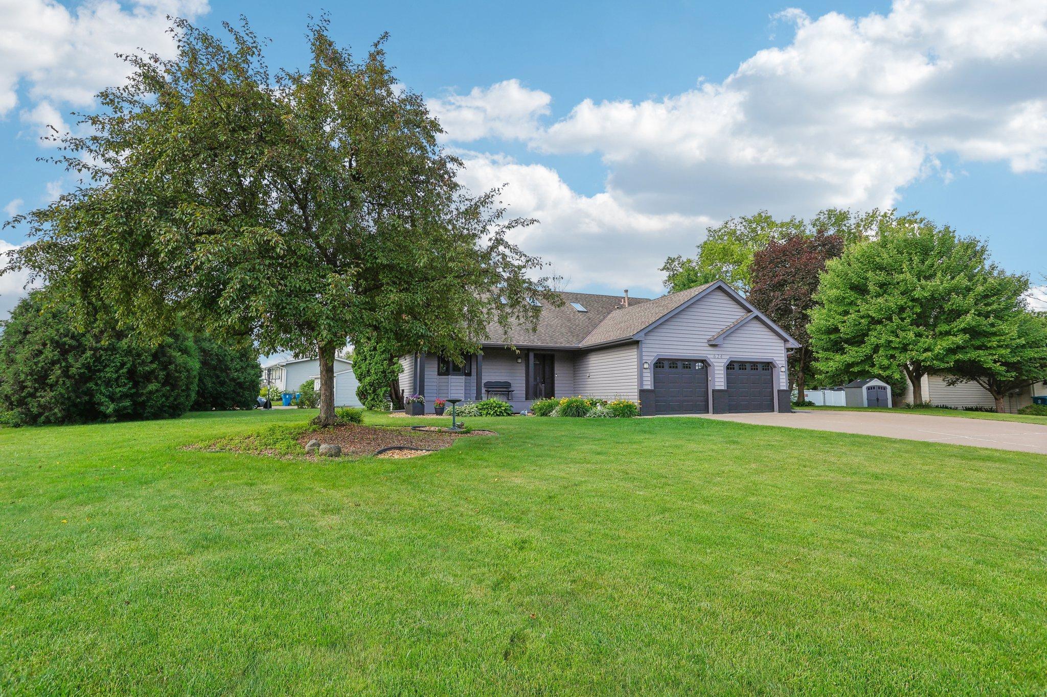 Gray single-story home with a two-car garage, broad green lawn, and mature trees.
