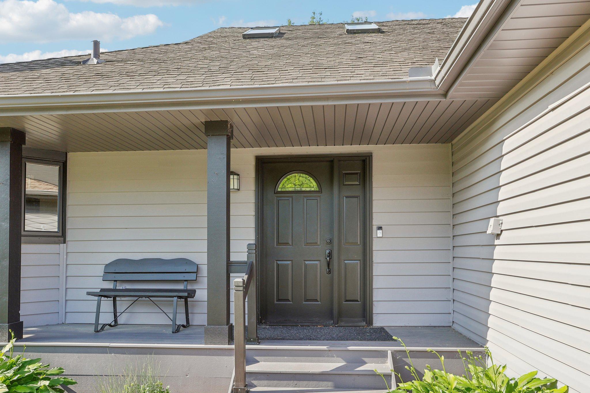 Covered front porch with a dark paneled door, gray siding, and a bench beside the entry.