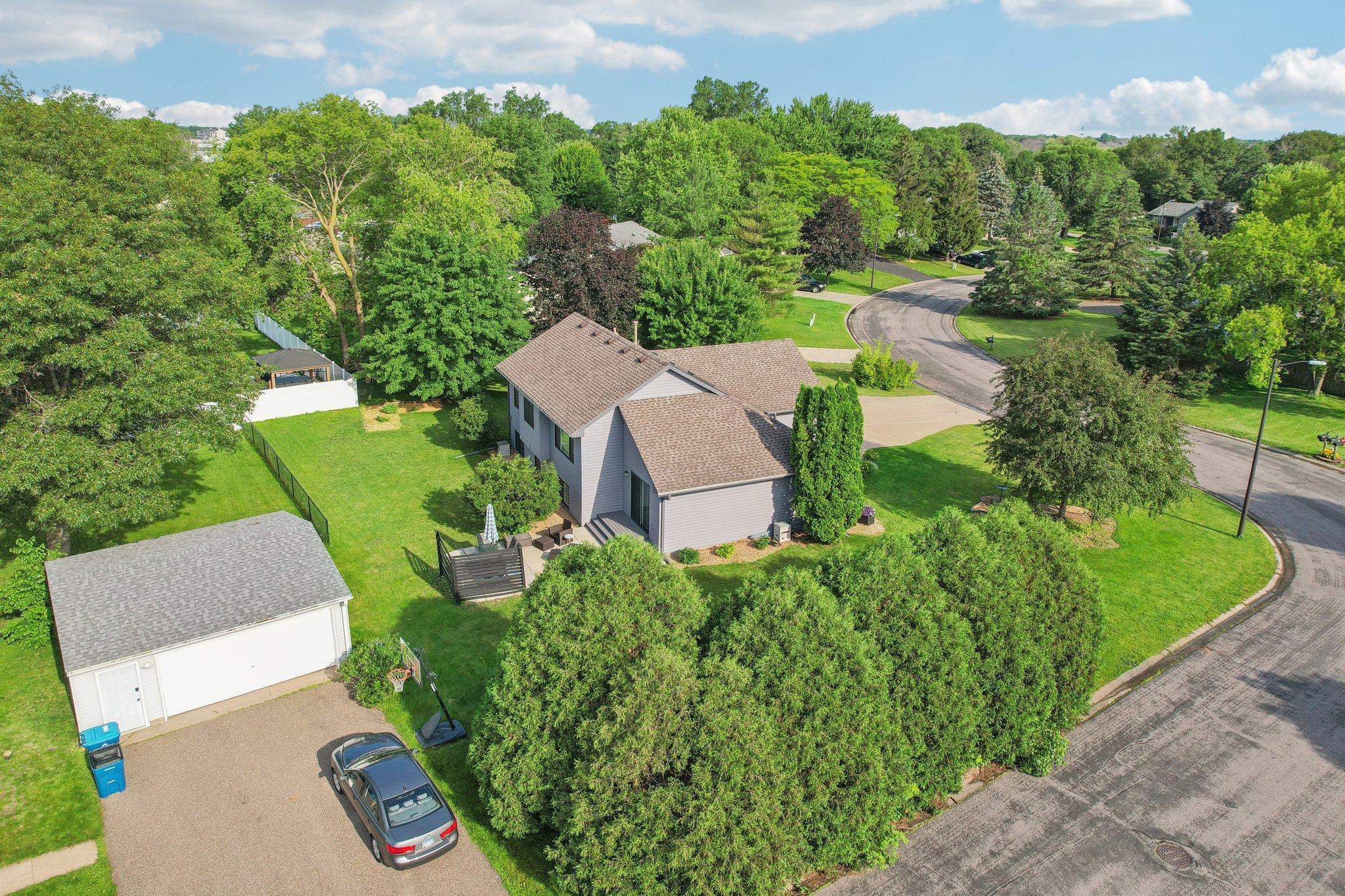 Aerial view of a gray-sided house with a detached garage, fenced lawn, and driveway beside a curved street.