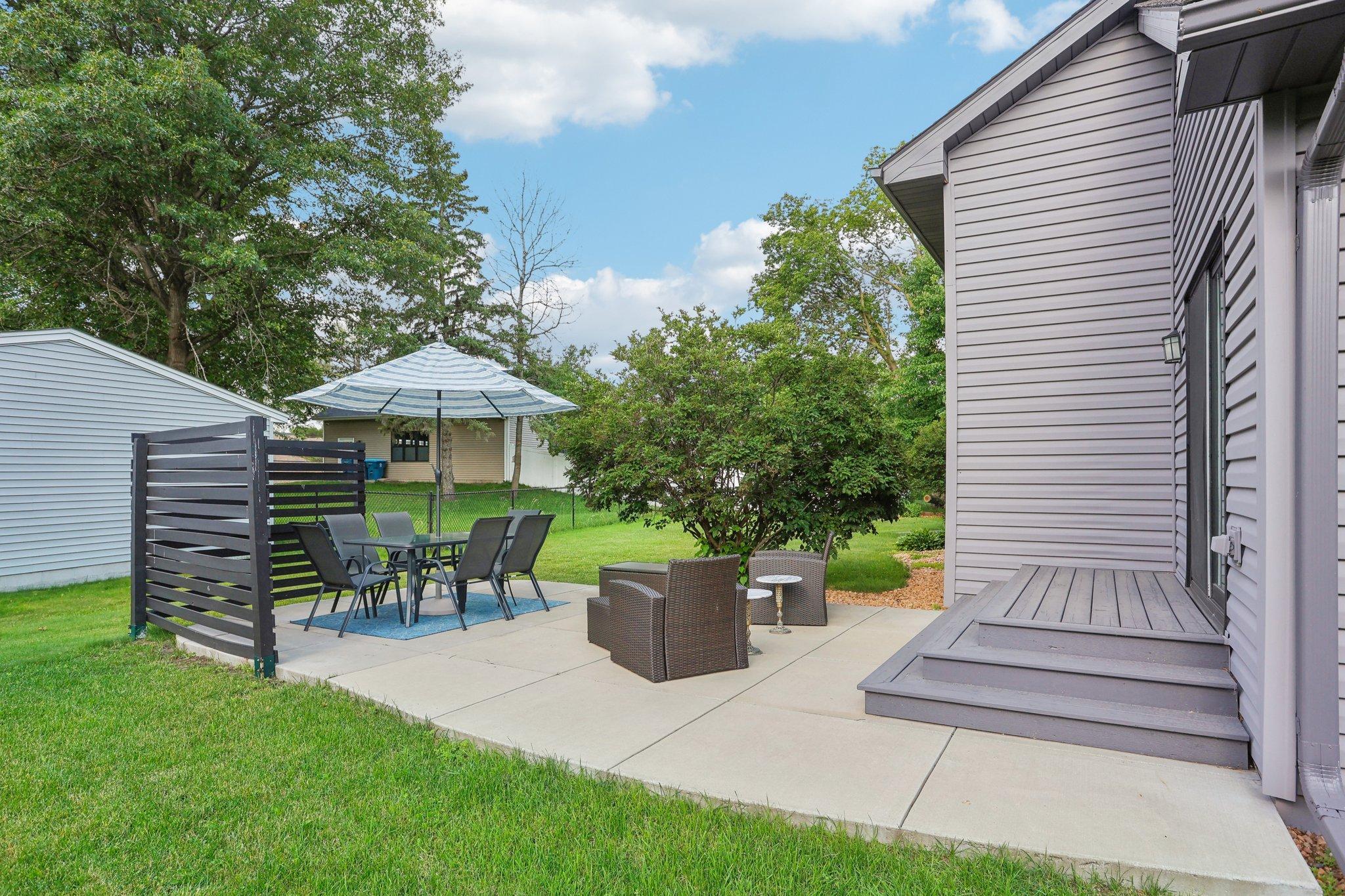 Back patio with outdoor dining beneath a striped umbrella and wicker lounge chairs beside the house.