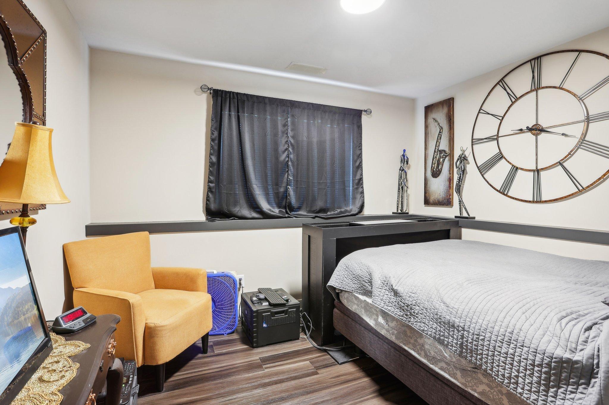 Furnished bedroom with a dark platform bed, mustard armchair, and oversized Roman numeral wall clock.