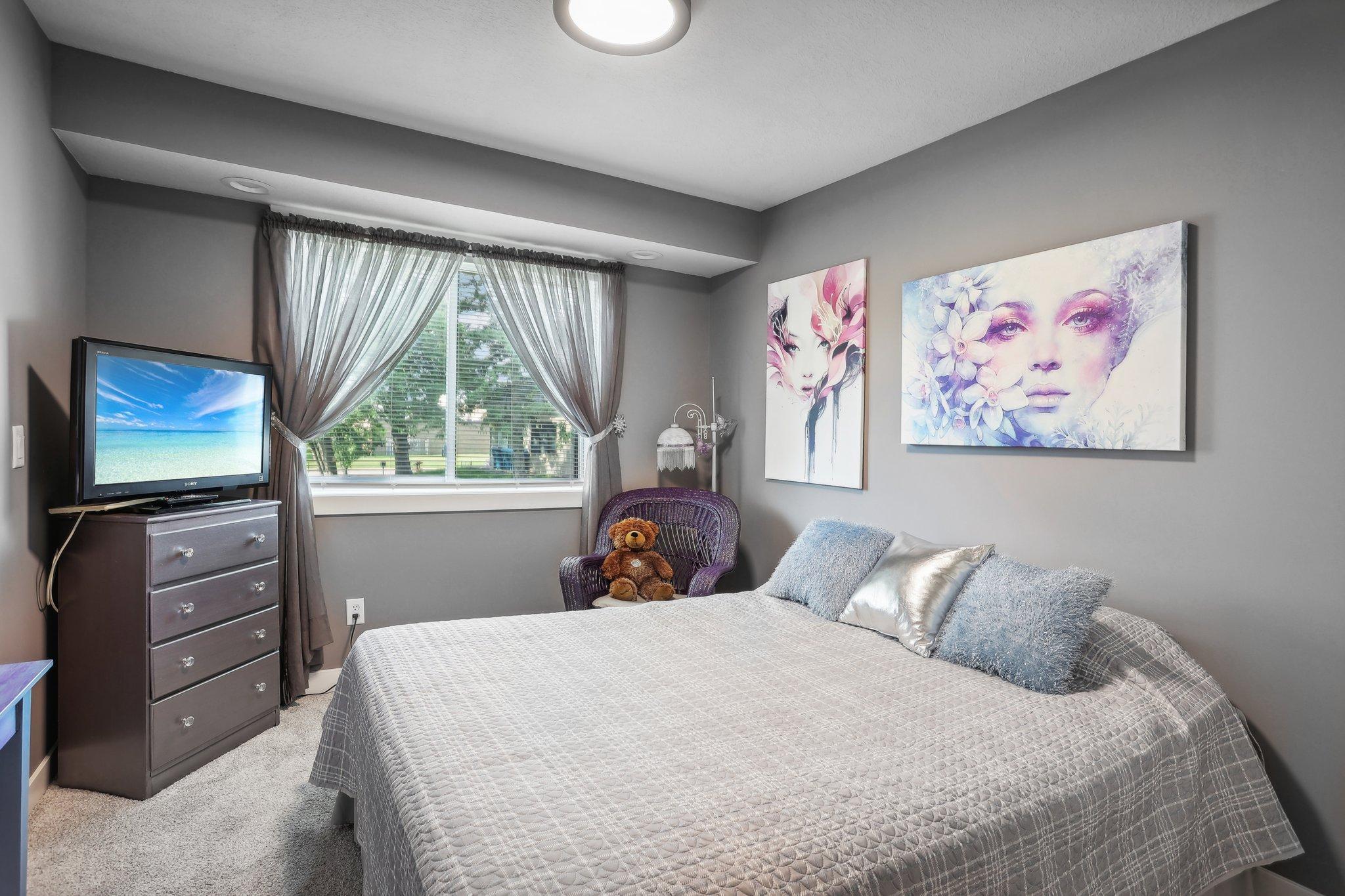 Furnished bedroom with a gray quilted bed, floral wall art, and a teddy bear in a wicker chair.