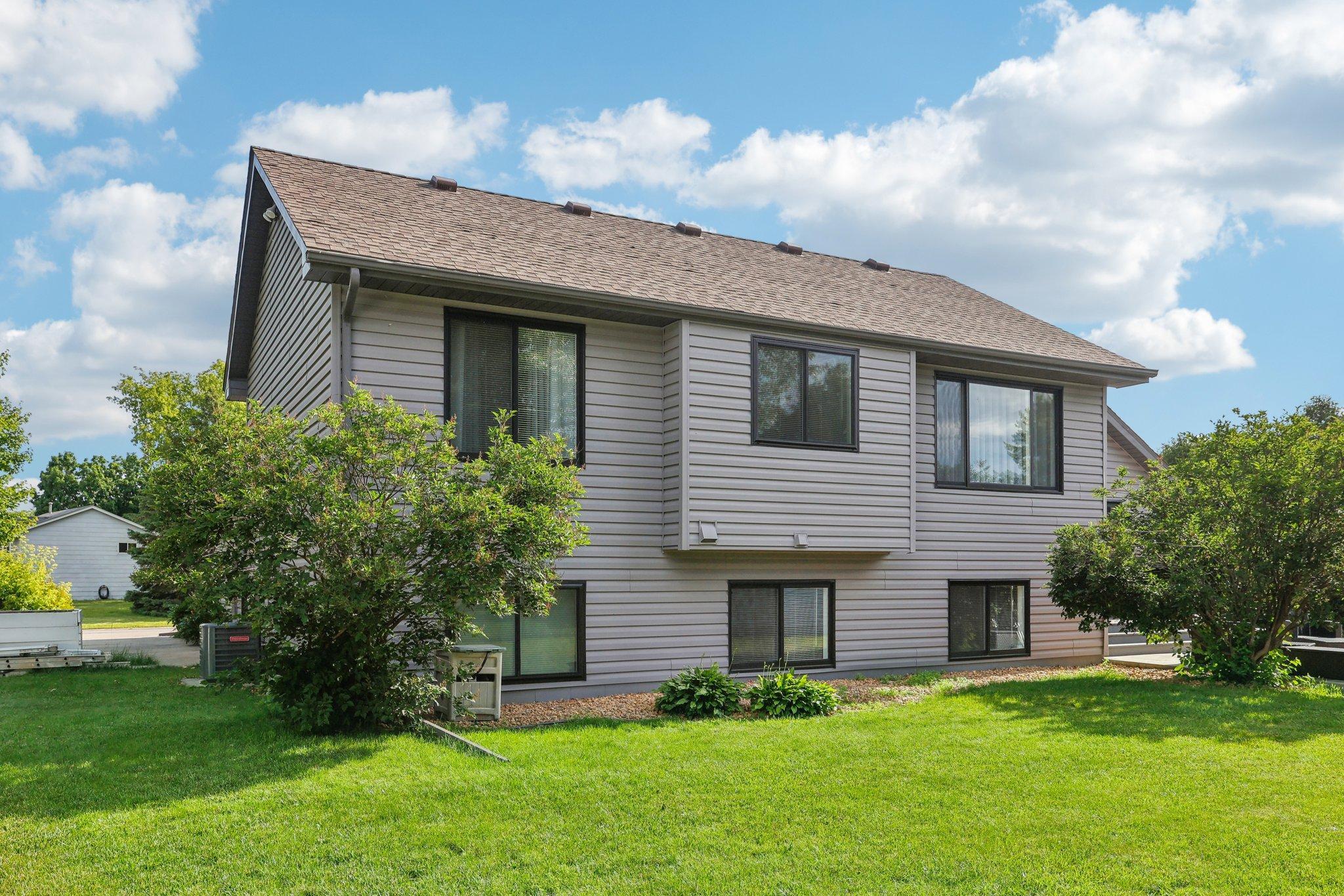 Gray two-story house with dark-framed windows, leafy shrubs, and a broad green lawn.