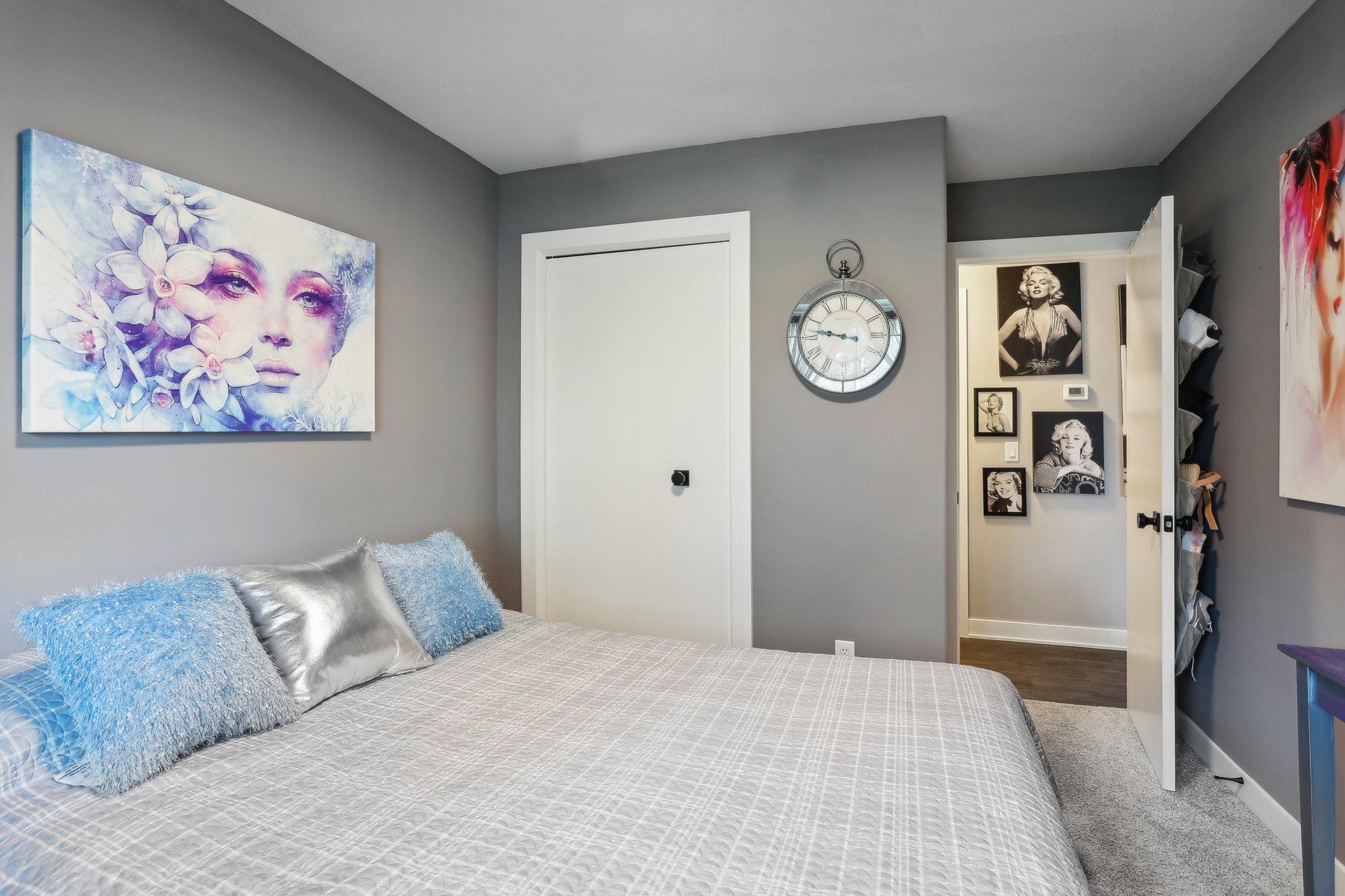 Furnished bedroom with blue pillows, colorful floral wall art, and an open doorway to a portrait-lined hall.