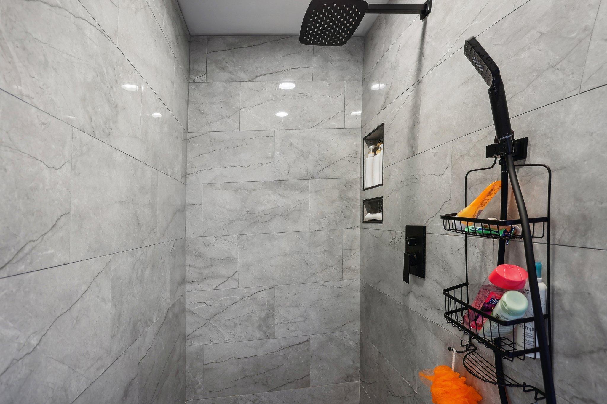 Walk-in shower with gray marble-look tile, black fixtures, recessed shelves, and toiletry baskets.