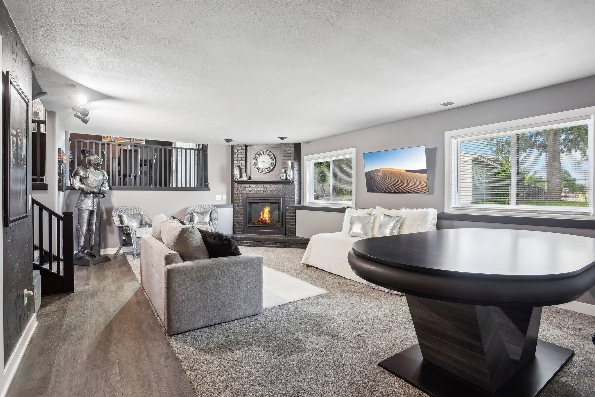 Furnished living room with a brick fireplace, oversized clock, and decorative suit of armor beside the stairs.