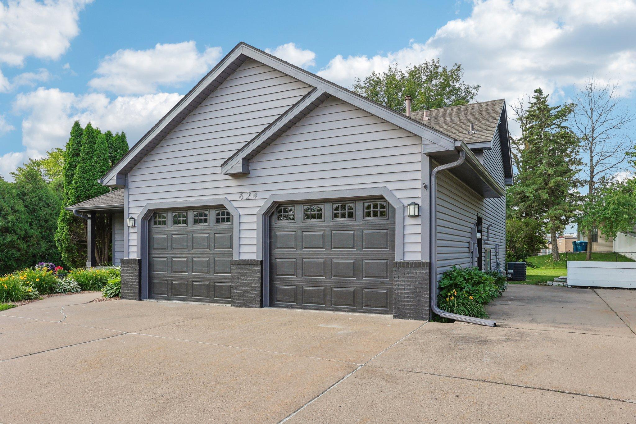 Gray house with two dark garage doors, a wide concrete driveway, and landscaped flower beds.