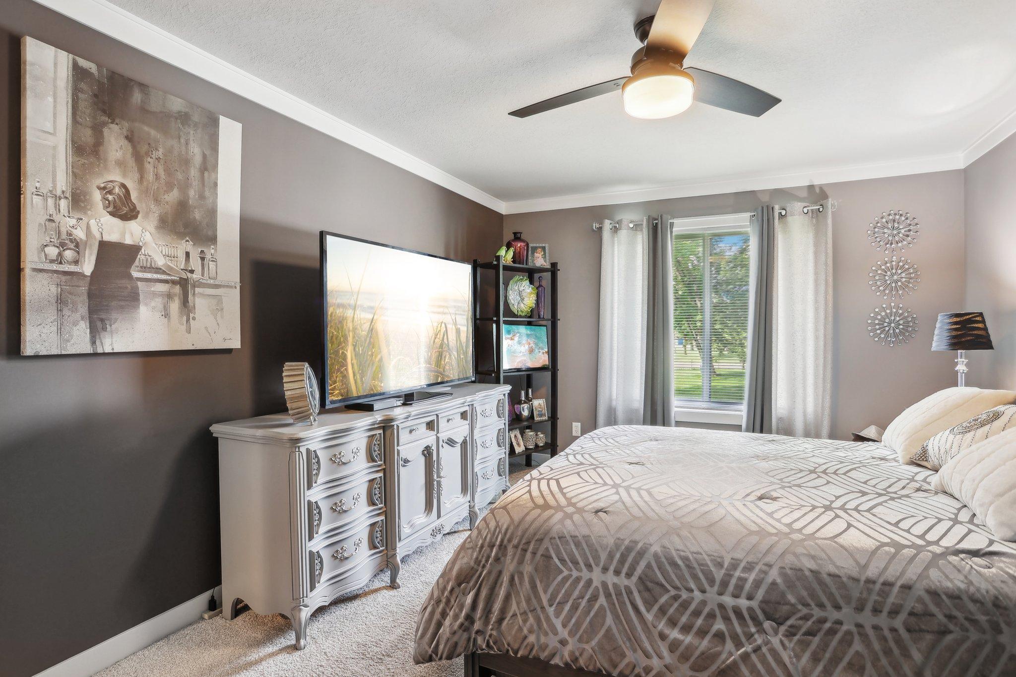 Furnished bedroom with a patterned bed, ornate white dresser, television, and window with gray curtains.