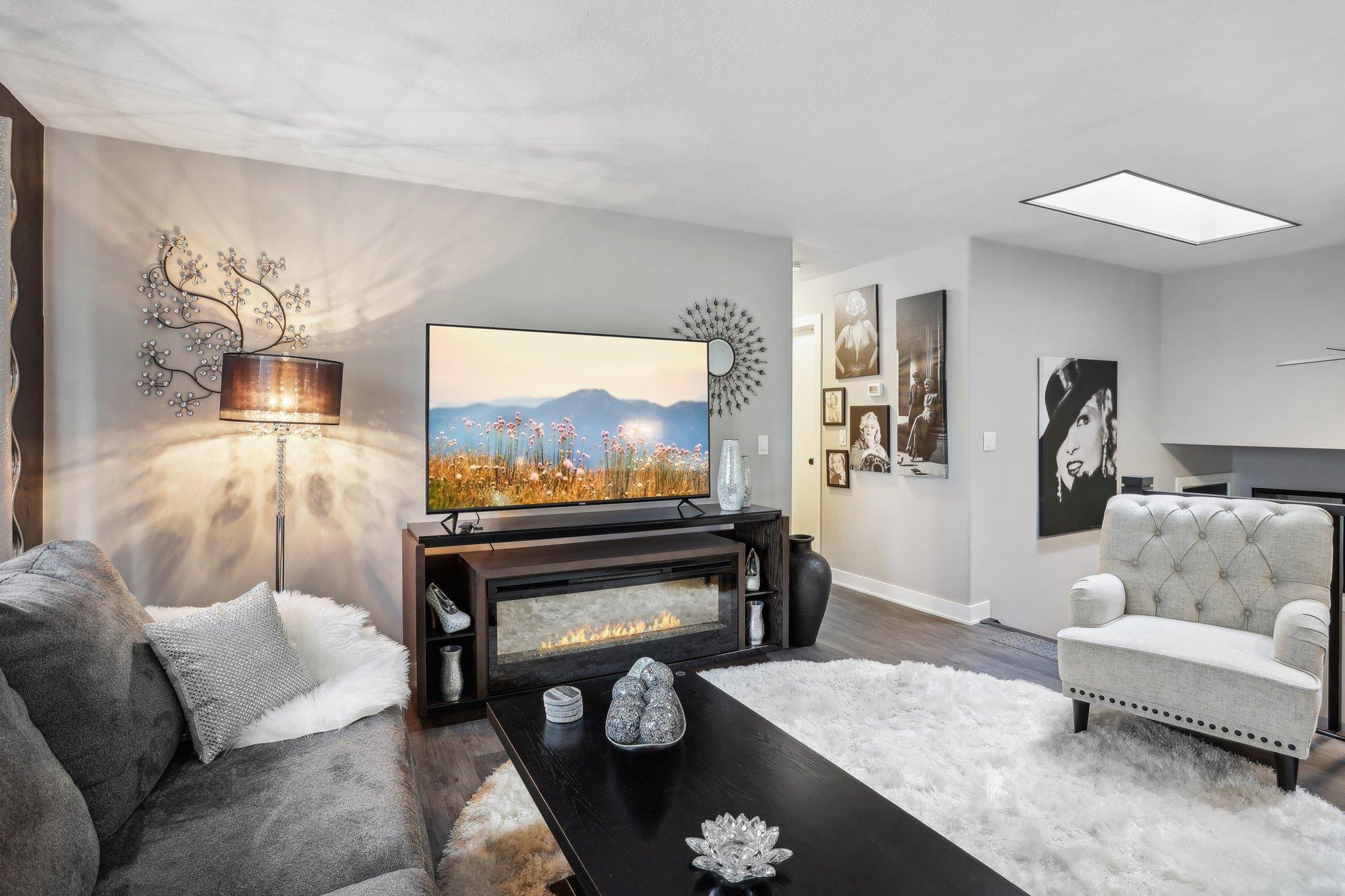Living room with a television above an electric fireplace, gray seating, and white shag rugs.