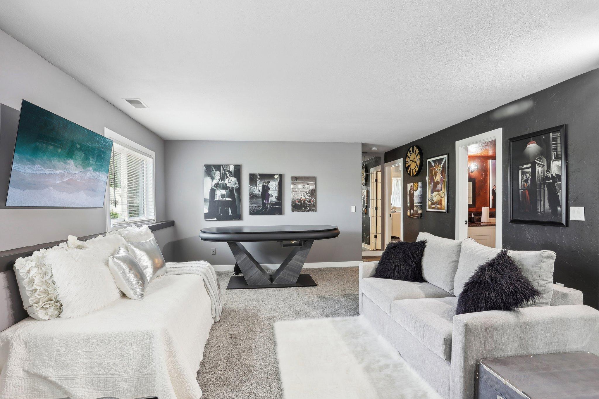 Living room with light sofas, an oval game table, wall-mounted TV, and framed artwork.