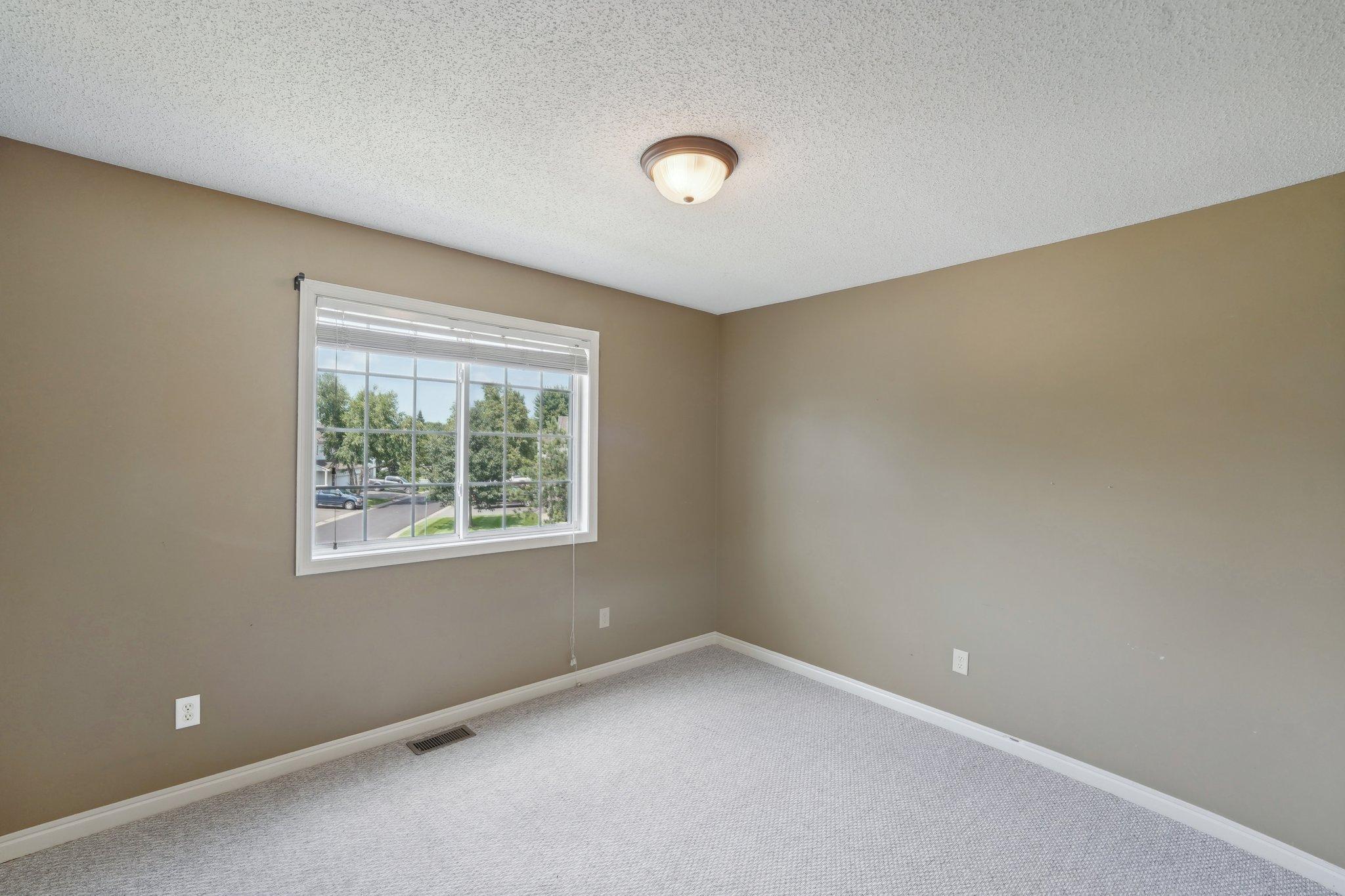 Empty carpeted room with taupe walls and a wide window overlooking trees and a residential street.