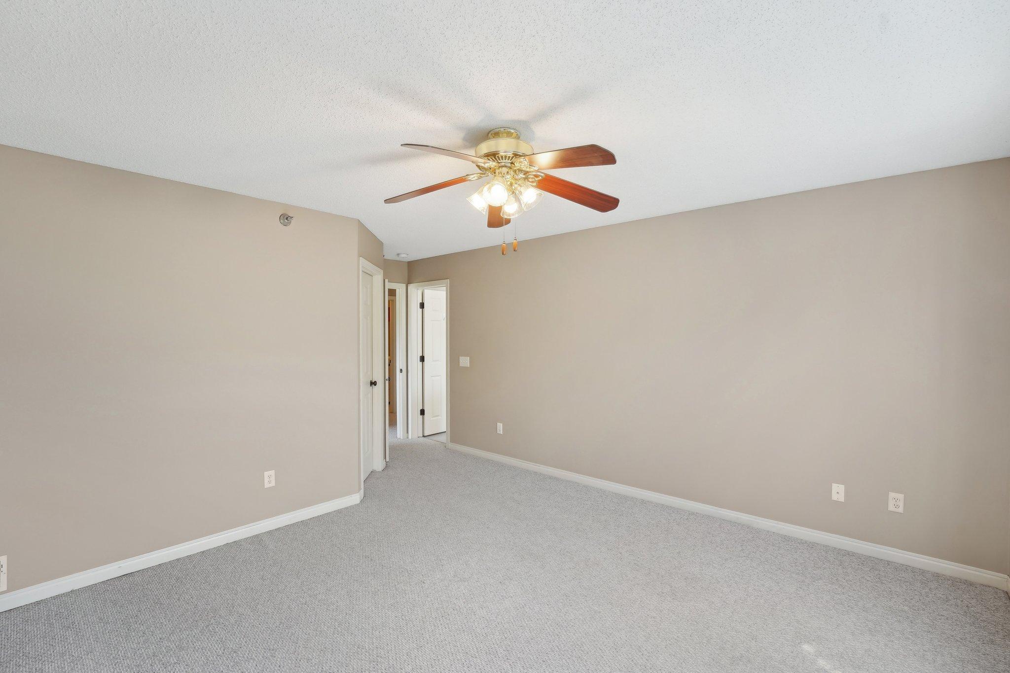 Empty bedroom with gray carpet, beige walls, and a ceiling fan with lights.