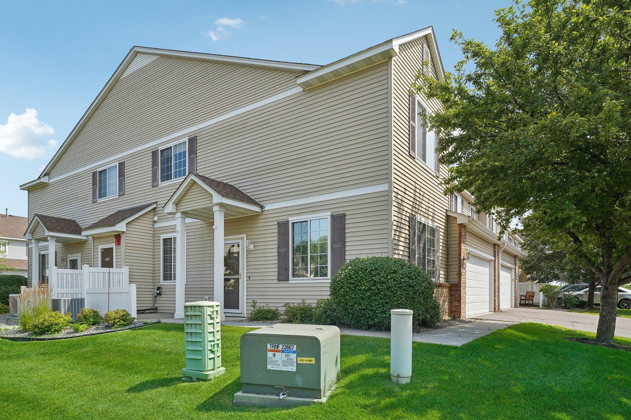 Beige two-story townhouse exterior with a covered porch, attached garages, and a large shade tree.