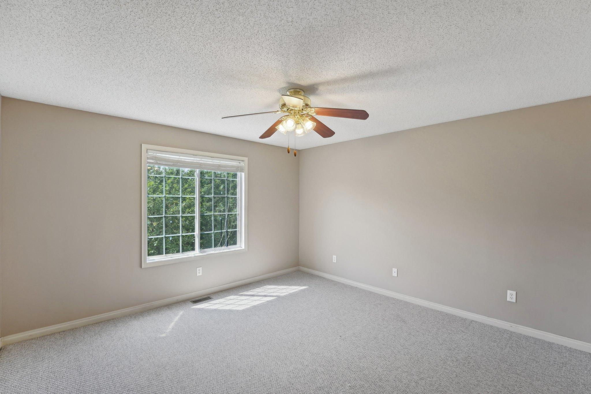 Empty carpeted bedroom with a large window and a lit ceiling fan.