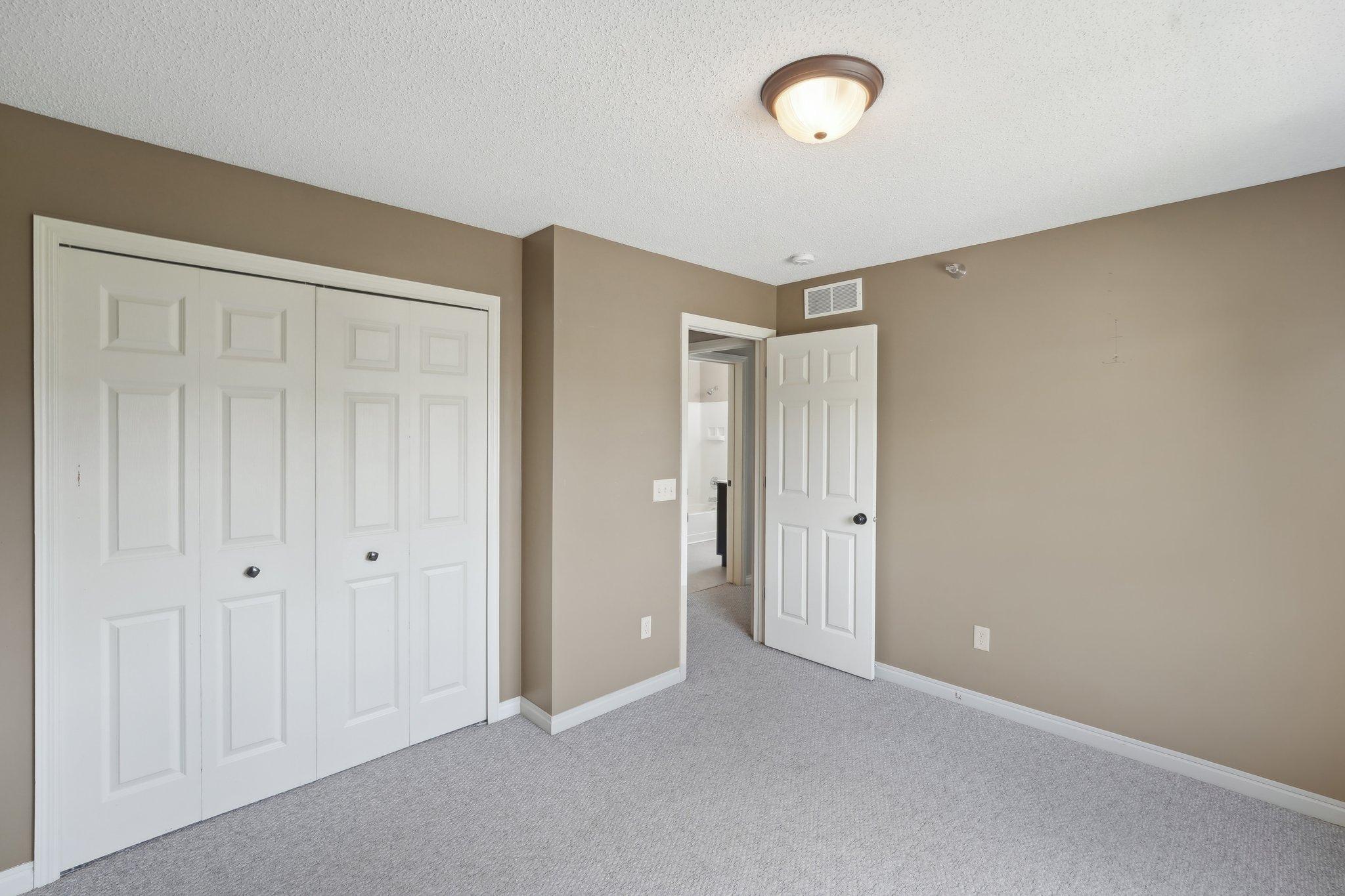 Empty bedroom with gray carpet, white closet doors, and an open bathroom door.