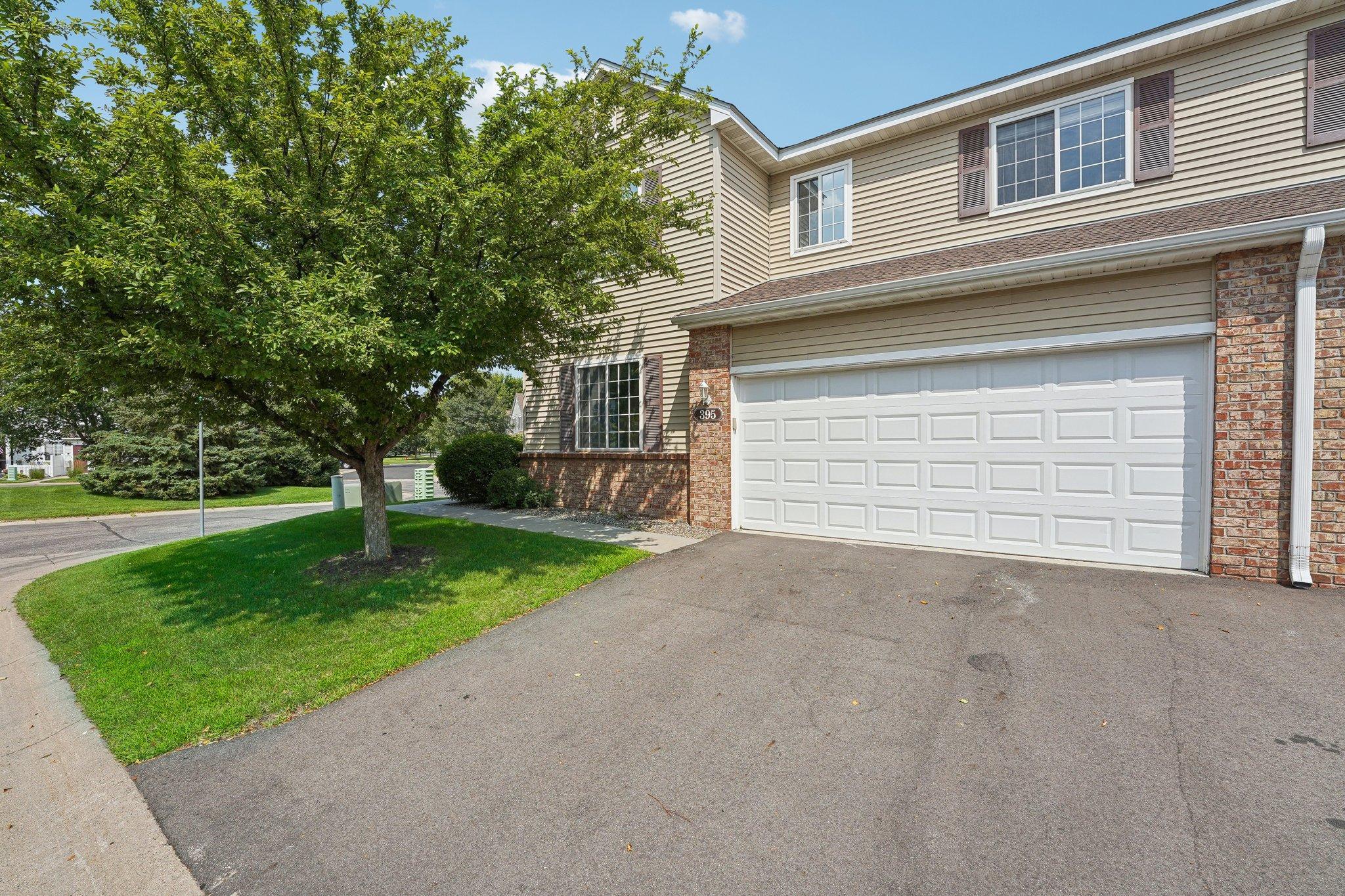 Two-story beige home with brick accents, a double garage, and a mature tree by the driveway.