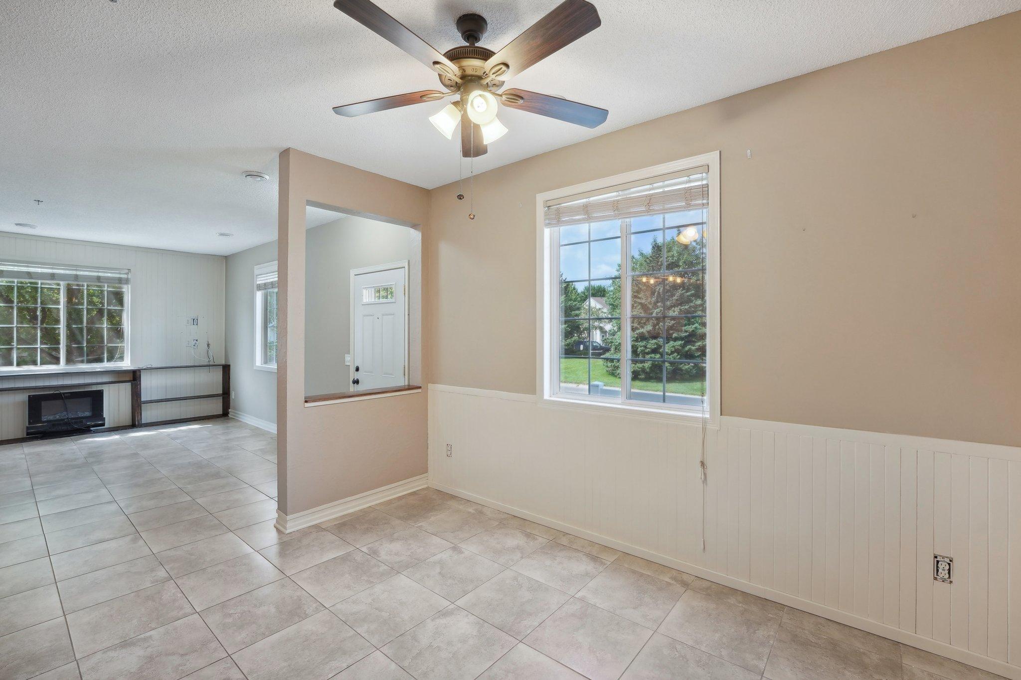 Empty tiled room with a ceiling fan, large window, and adjoining fireplace area.