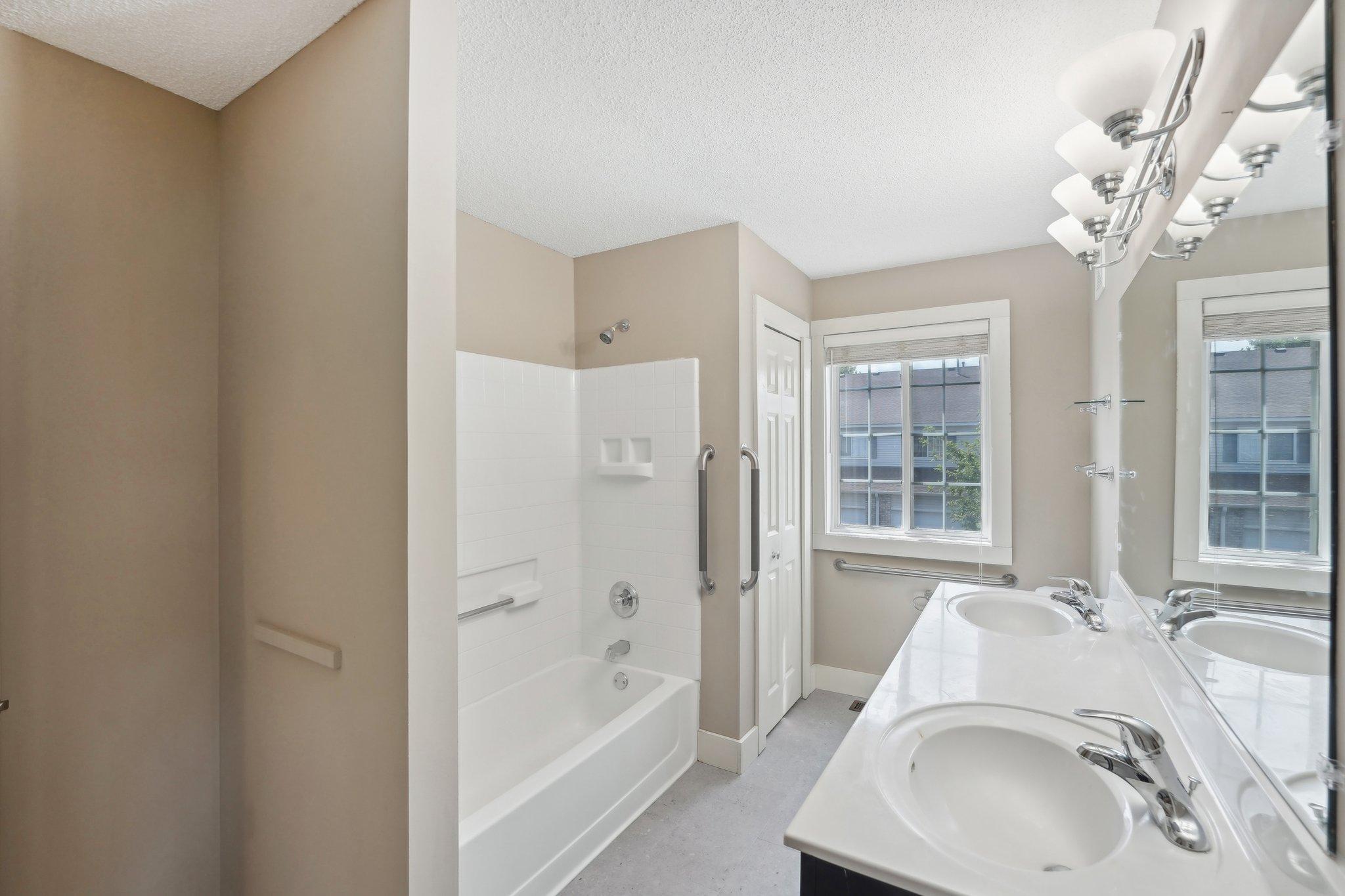 Bathroom with a white tub and shower, grab bars, and a double-sink vanity beside a window.