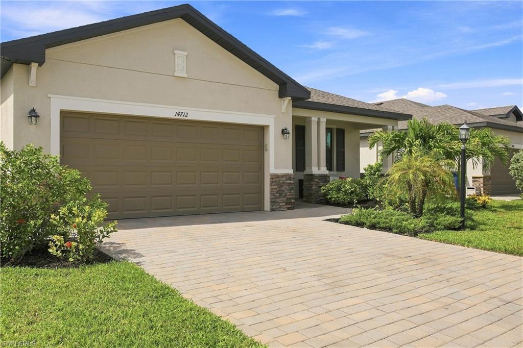 Beige single-story home with a two-car garage, paver driveway, and tropical landscaping.