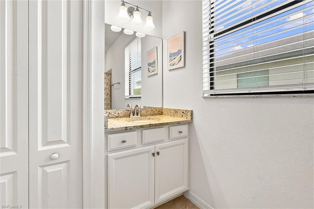 Bathroom vanity with white cabinetry, speckled granite counter, large mirror, and a window with blinds.