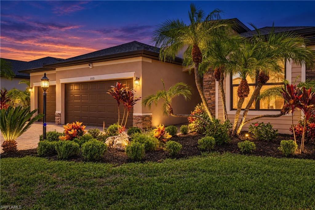 Home exterior at dusk with a two-car garage, palm trees, and colorful tropical landscaping.