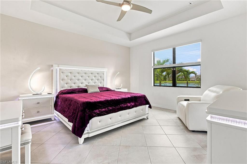 Furnished bedroom with a white tufted bed, plum coverlet, and window overlooking water and palm trees.