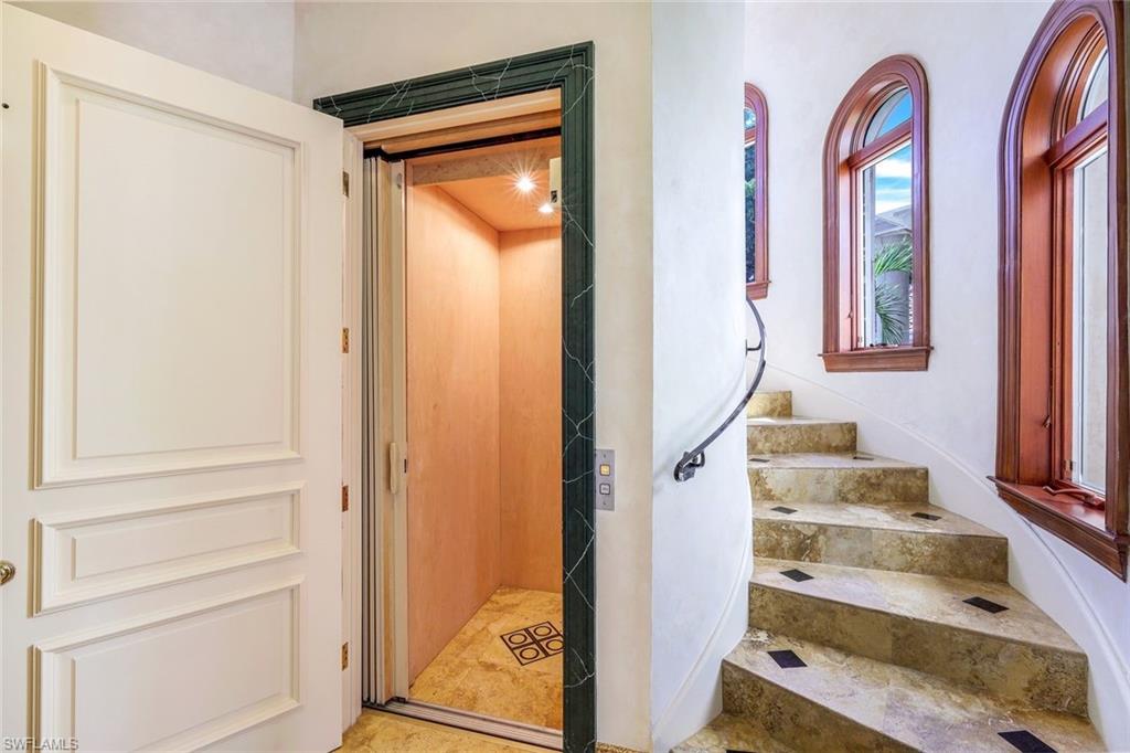 Open elevator beside a curved stone staircase with arched wood-framed windows.