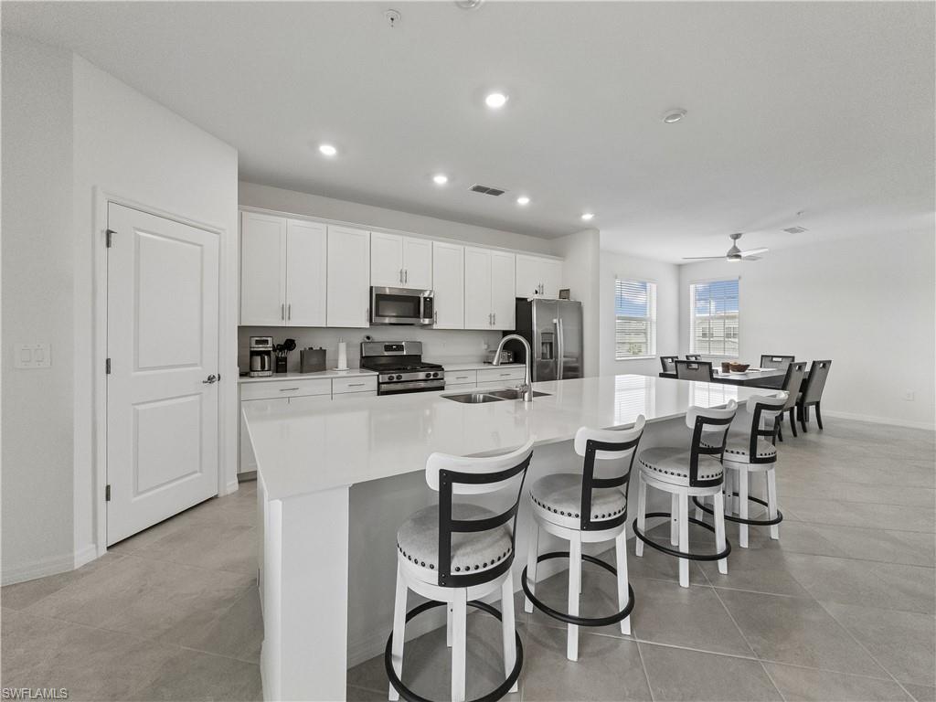 Modern white kitchen with a large island, five stools, stainless appliances, and an adjoining dining area.