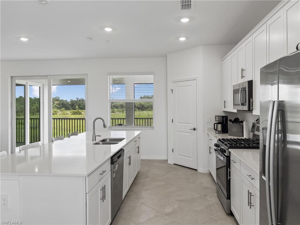 Modern white kitchen with a large island and stainless steel appliances, overlooking a green field.