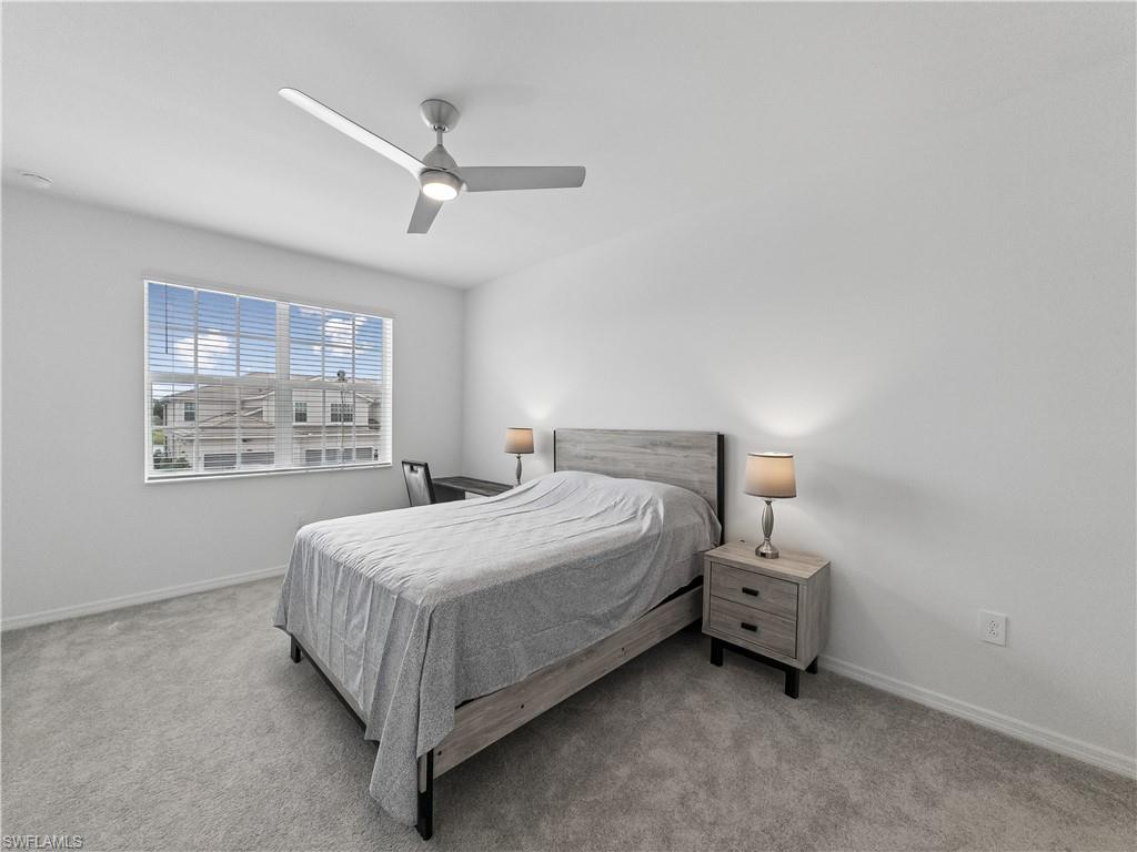Staged bedroom with a gray bed, matching nightstand, ceiling fan, and window overlooking neighboring homes.