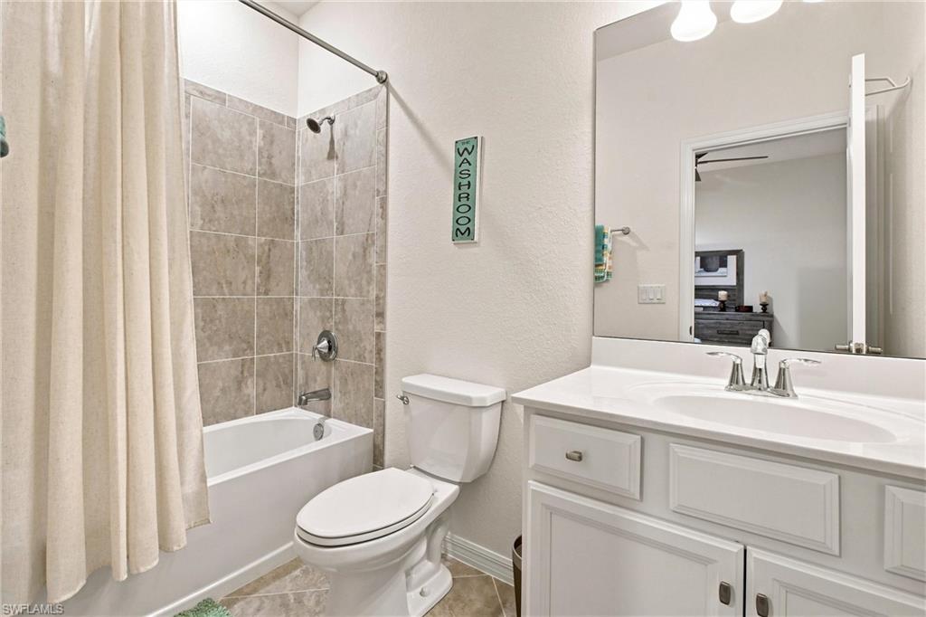 Bathroom with a tiled tub and shower, cream curtain, toilet, and white vanity.