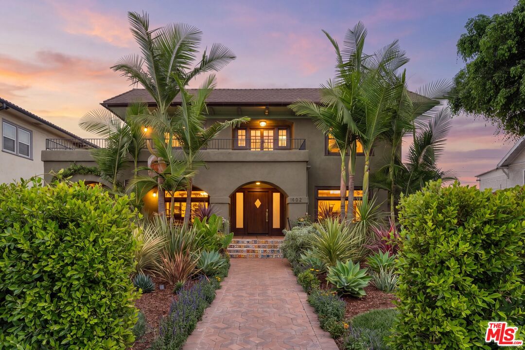 Two-story home with a warmly lit entry, palm trees, and a tiled front walkway at dusk.