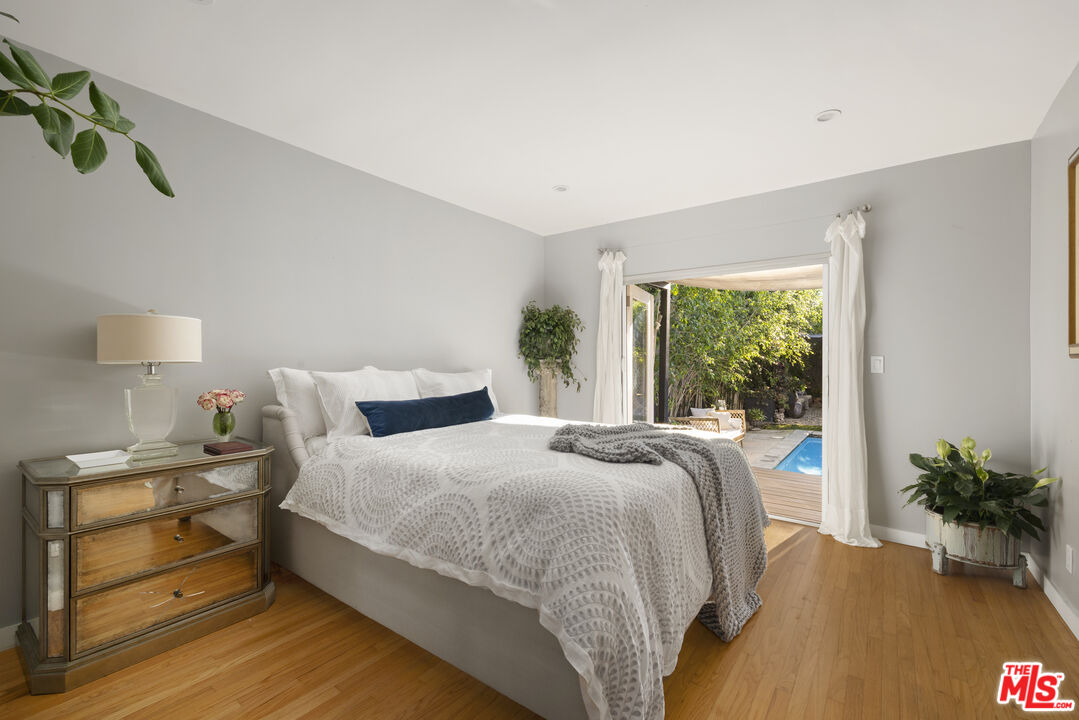Staged bedroom with a white bed, hardwood floors, and open doors overlooking a pool.