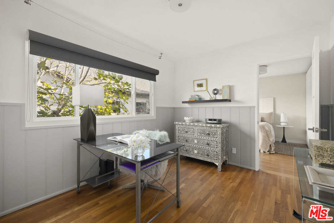 Furnished home office with a glass desk, patterned dresser, and large window overlooking leafy branches.
