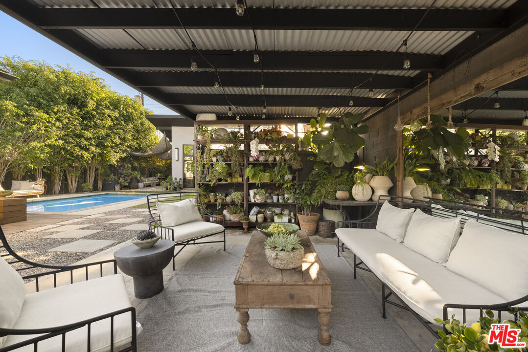 Covered patio with white-cushioned seating, abundant potted plants, and a swimming pool.