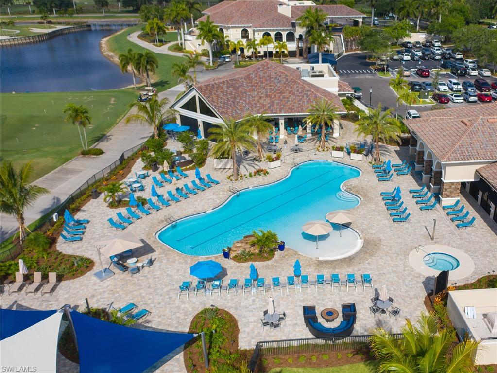 Community pool and spa surrounded by lounge chairs, palm trees, clubhouse buildings, and a lakeside lawn.