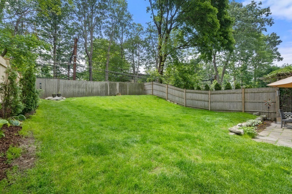 Fenced backyard with a broad lawn, mature trees, and a small patio.