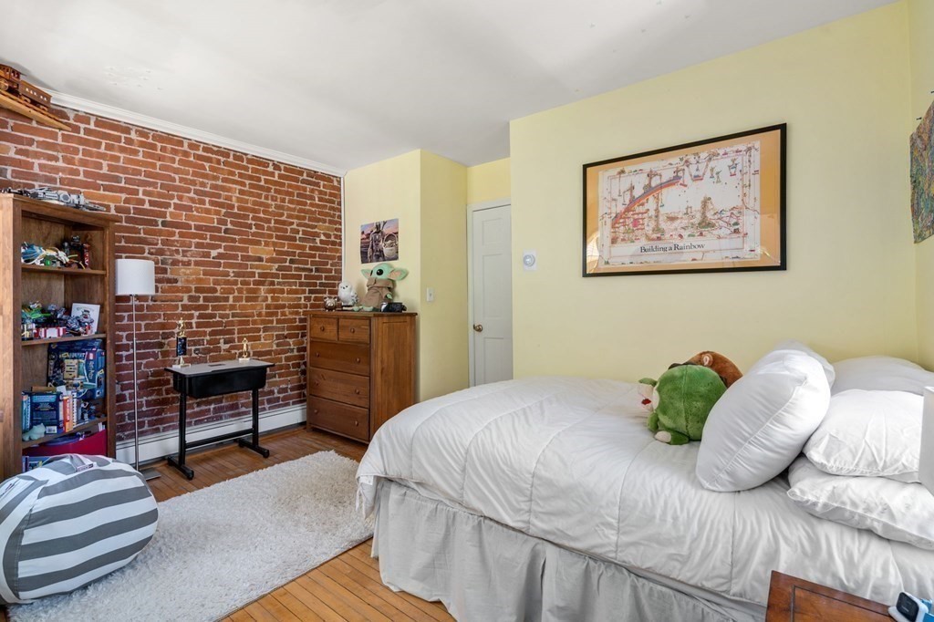 Furnished bedroom with a white bed, exposed brick wall, and hardwood floors.